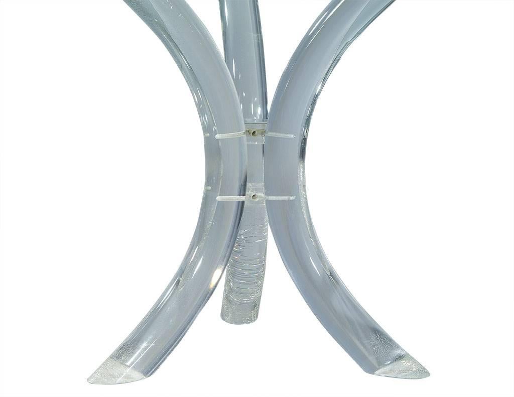 A Classic Mid-Century Modern accent piece, this table features a grand glass top with a thick section and wide bevelled edge, resting on a tripod made up of three heavy, curved Lucite members forming a saber legged pedestal, joined at the centre