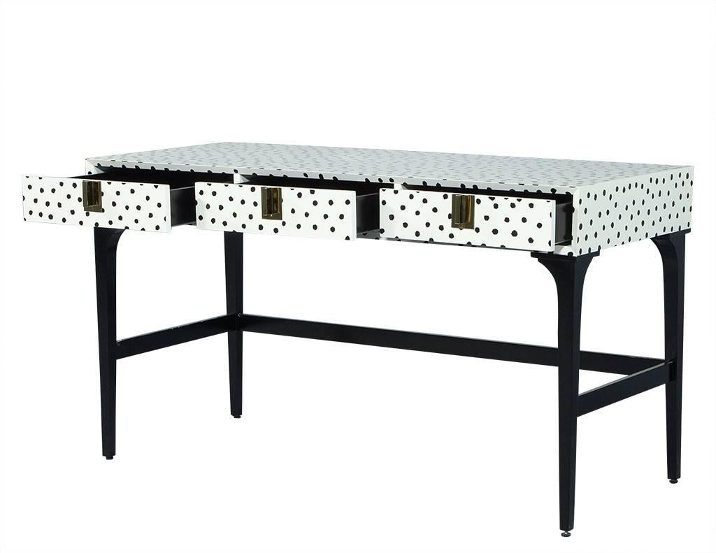This modern desk contains three drawers and is accented with polished brass square pulls. The piece sits atop four tapered black legs connected by stretchers, and the top is finished in a creamy white lacquer covered in hand-painted black polka