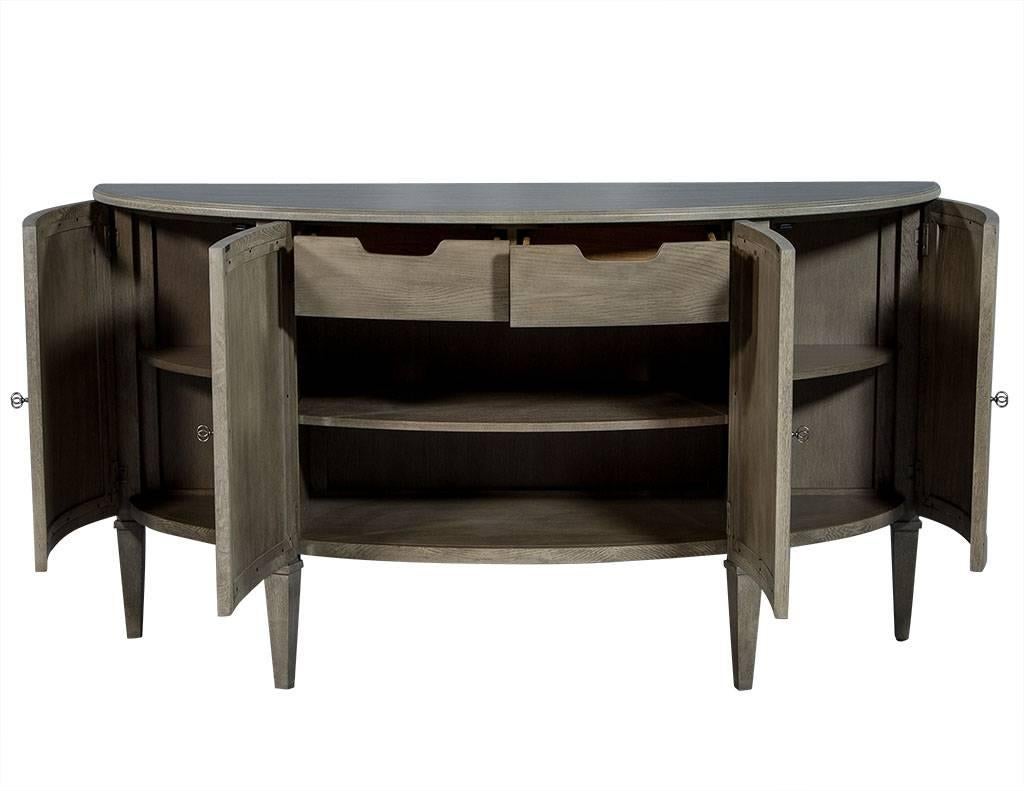 This traditional style sideboard is designed by Bernhardt. It is composed of oak veneers in a heather grey finish with four cabinet doors all atop four fluted legs. The demilune shape and inlay on the front exterior adds depth and character, while