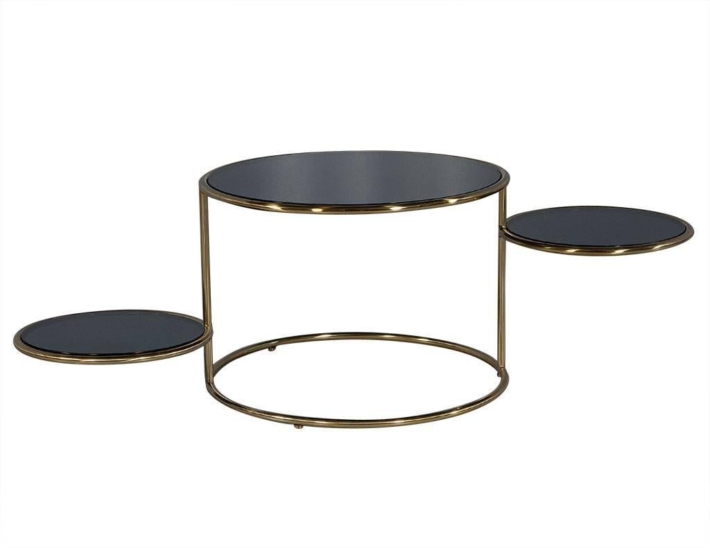 This modern cocktail table is richly designed. It is crafted out of a brushed brass frame with a black glass top and includes two smaller rounded black glass shelves that can swivel under the top or on the exterior of the piece. A unique and