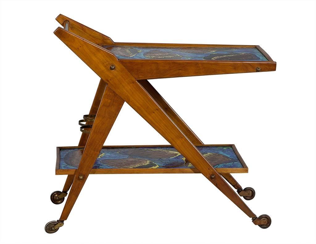 This Art Nouveau style serving cart is an actual work of art! Crafted out of mahogany with an original signed oyster motif on the ceramic inlays of both tiers, it sits atop casters for ultimate mobility. For added utility, it has a metal three ring