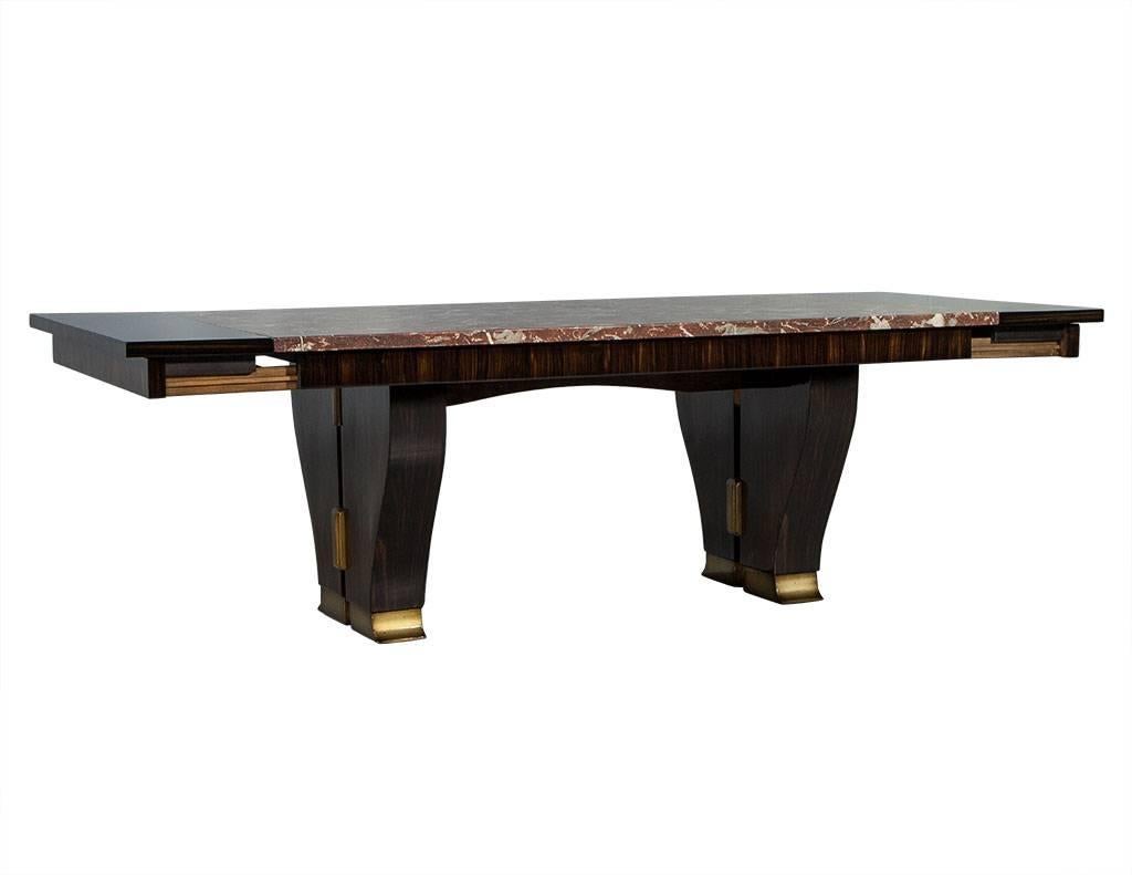 This Art Deco style table is rich and bold in design. It showcases the original burgundy marble top and has two wood extensions for each end. The Macassar wood double pedestal is tapered yet dense and the feet are painted with gold leaf. A Carrocel