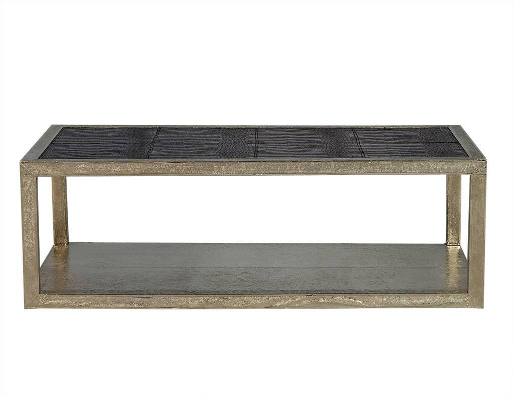 This modern cocktail table is metal encased and covered in a black faux crocodile leather finish on the top surface. It has distressed, silver detail on the lower level, legs, and edge of the top surface. A gorgeously polished piece perfect for a