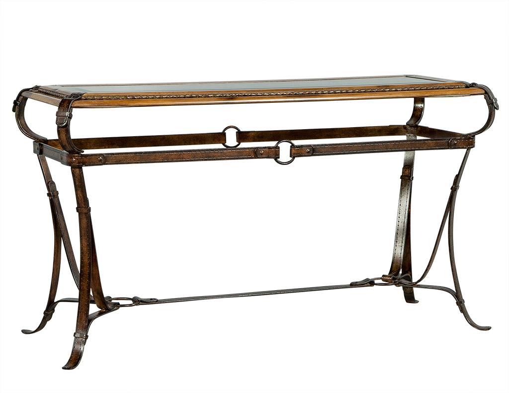 This Rancho Monterey Style console table is ornate and spectacular. It is composed of light walnut-finished wood with inset beveled glass on top. The glass is framed with a leather braid trim and the four-footed base is composed of a wrought iron