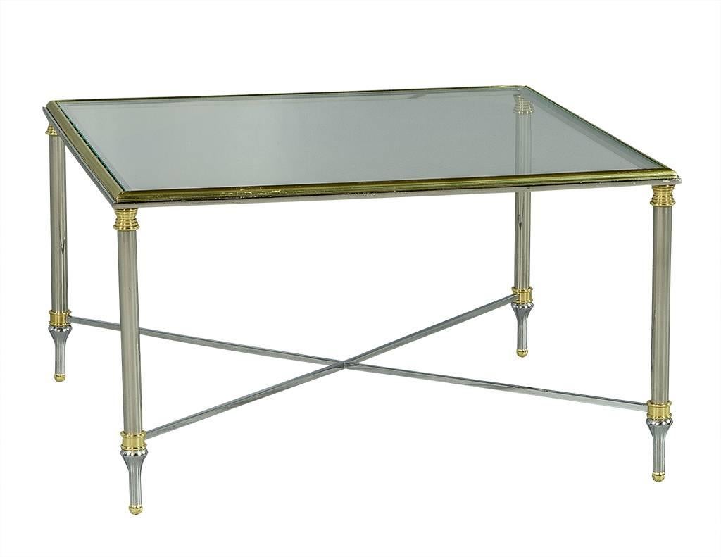 This stylish Maison Jansen inspired cocktail table has an X-base chrome frame and inset square glass top. Adorned with brass accents, and delicate fluted legs, this table is a great fit for a timeless living space.