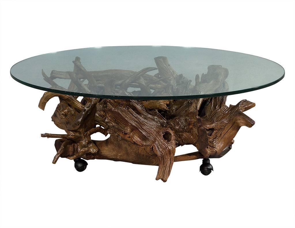 This rustic cocktail table is full of character. It is comprised of a glass top with a wood sculpture base on casters. Perfect for a bold and inviting home.