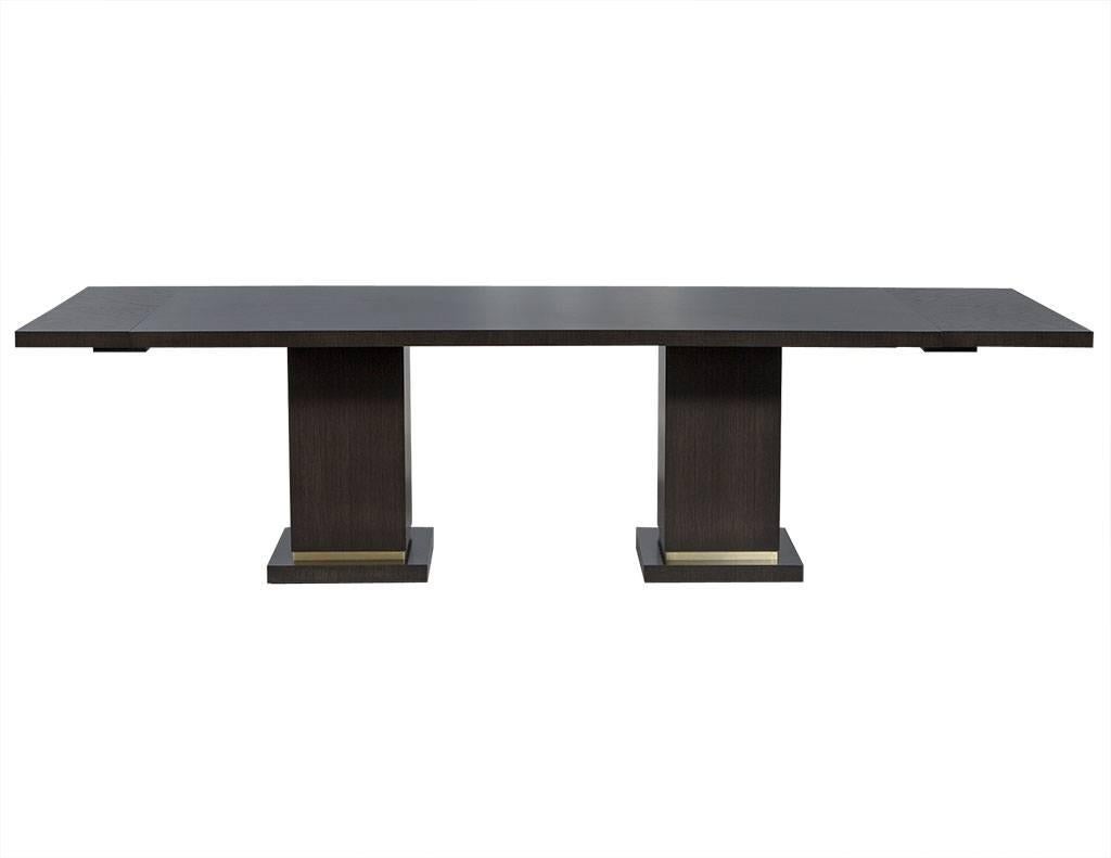 This modern dining table is a beautiful custom Carrocel piece. Brand new, it is crafted out of solid oak and has two leaves of different widths for each end. The piece sits atop two square pedestal bases with gold metal trim at the bottom. A perfect