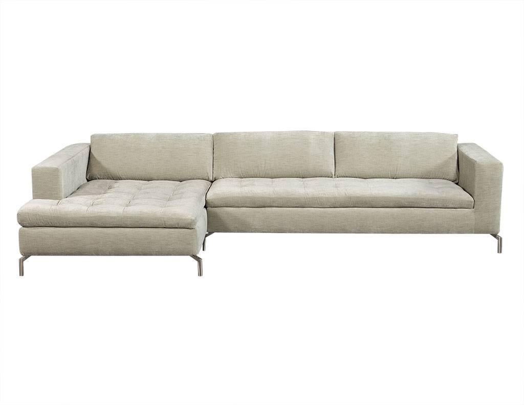 This modern sectional sofa is wholly pristine. It has an extended chaise section and sofa section, and is upholstered in a light grey fabric with attached pillow backrests. The seats are tufted and the upholstery is brand new. The piece sits atop