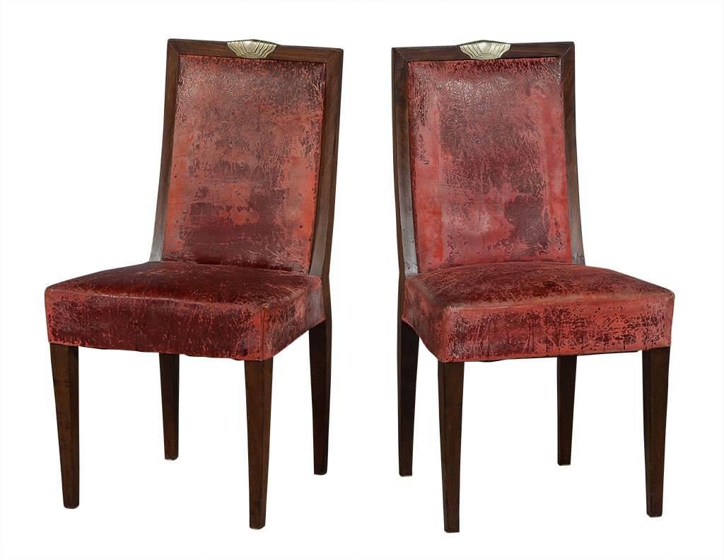 This set of six Art Deco style dining chairs hails from France. Each chair is comprised of a solid wood frame, distressed leather wraparound the edges, deep red upholstered seat, and sculpted metal detail on the top. In very good condition, the