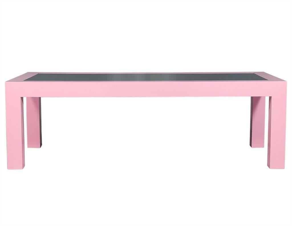 This modern, dining table is a Carrocel Custom piece. It is composed of solid wood finished in a pale pink with an inlaid dark glass top surface. Sitting atop large square legs made the same size as the wood on all four sides of the tabletop, this