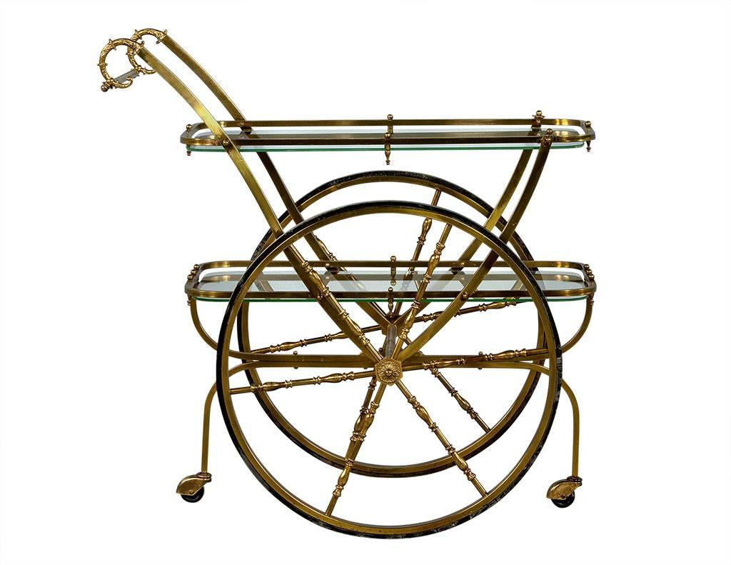 This bar cart is crafted out of brass and glass with large wheels at the sides and small caster wheels at the front and rear. The piece has brass detailing on the handles and wheel spokes, and is a perfect piece to add interest to a Mid-Century