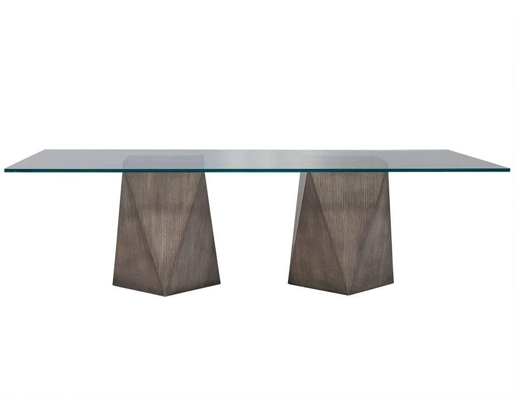 This ultra-cool dining table features a tempered glass tabletop with polished edges and sits atop two pedestals carved into a geometric eight-sided triangular design with a bleached grey finish. The piece can be ordered in custom sizes and different