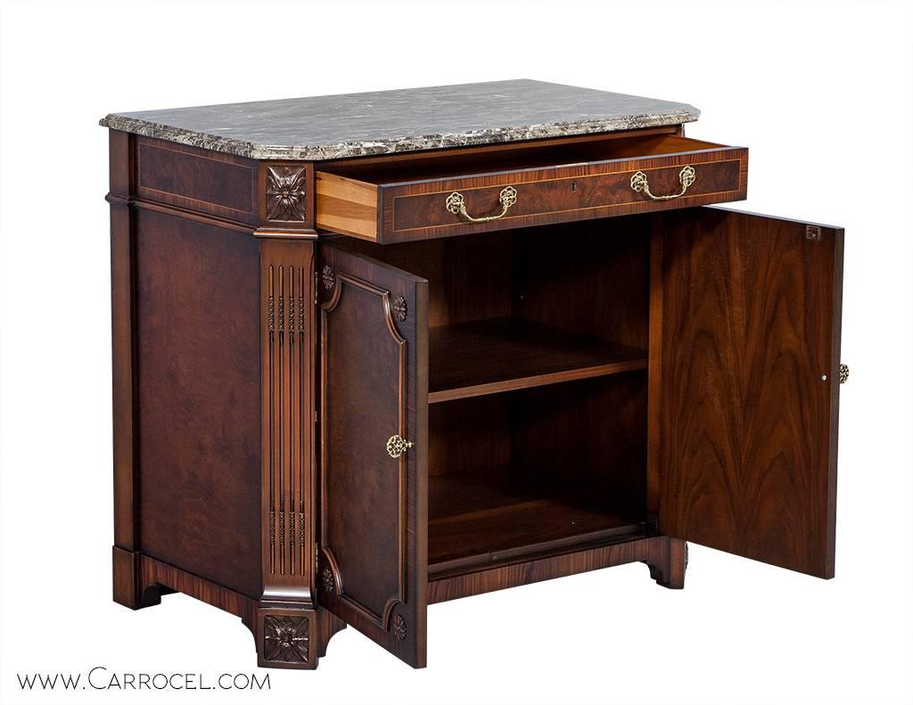 Marble top mahogany cabinet made with a beautiful sampling of veneer finished in rich mahogany tones. The façade is decorated in carved floral motifs with angled fluted columns framing each side and brass hardware. The cabinet is divided into a top