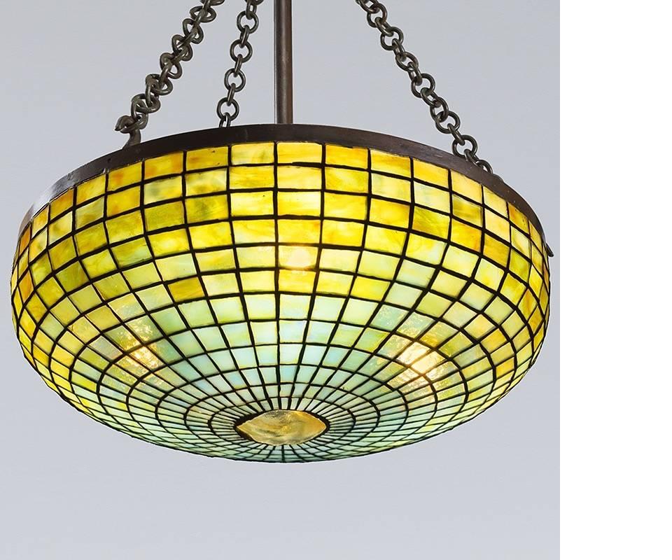 A Tiffany Studios New York glass and bronze “Geometric” chandelier, featuring a mottled yellow, cyan and orange leaded glass shade with a yellow/white iridescent glass “Turtleback” tile oculus center.

A similar chandelier is pictured in: Tiffany