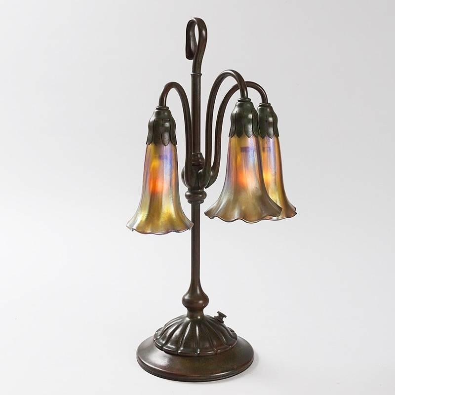 A Tiffany Studios New York glass and bronze three-light “Lily” desk lamp, featuring three golden iridescent Favrile “Lily” shades suspended from an decorated natural patina bronze base.

A similar lamp is pictured in: Tiffany Lamps and Metalware: An