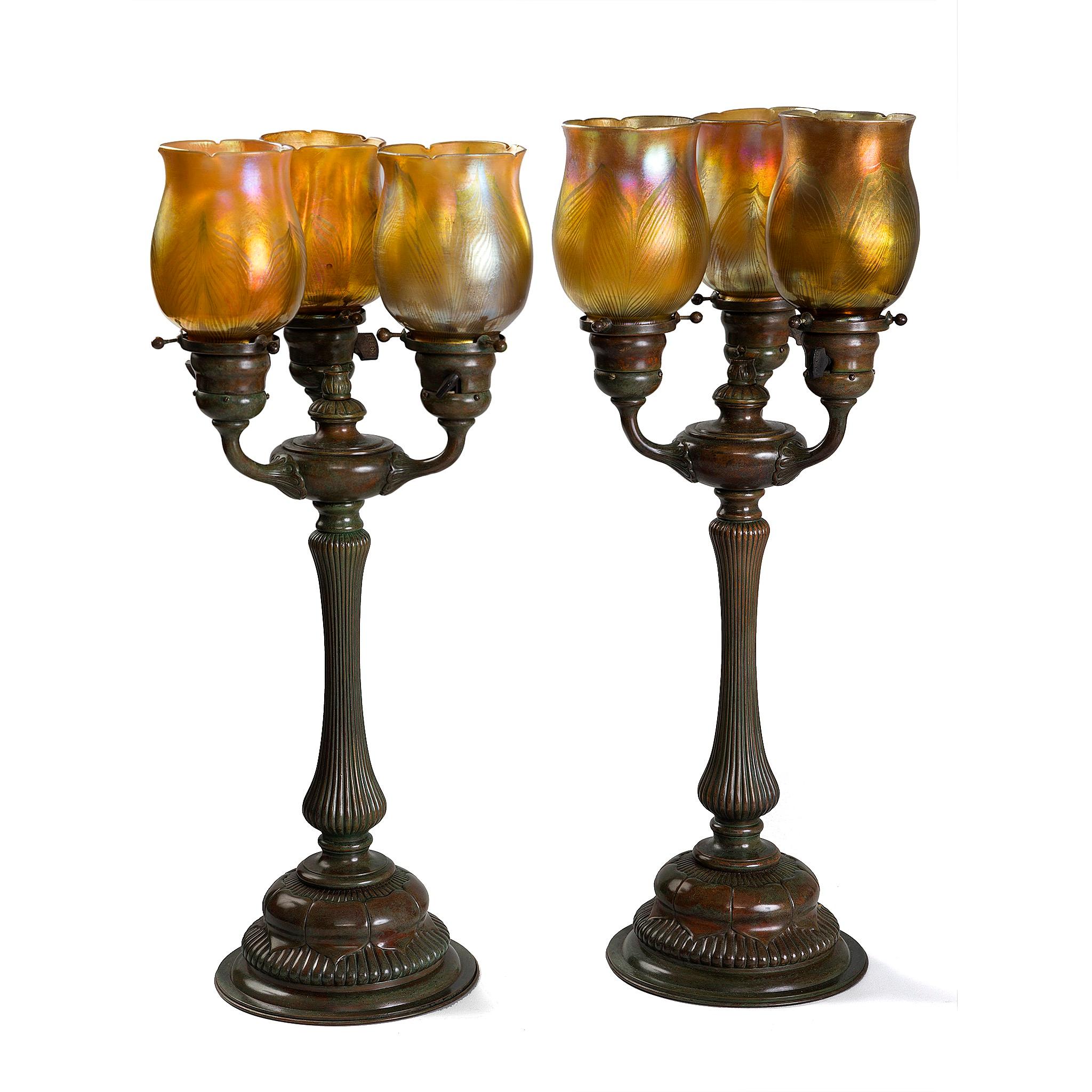 The Three-Light Lamp by Tiffany Studios New York showcases three gold Favrile glass shades with a subtle purple iridescence, elegantly mounted on a three-arm patinated bronze base. Inspired by the form of a candelabra, the design was intended to