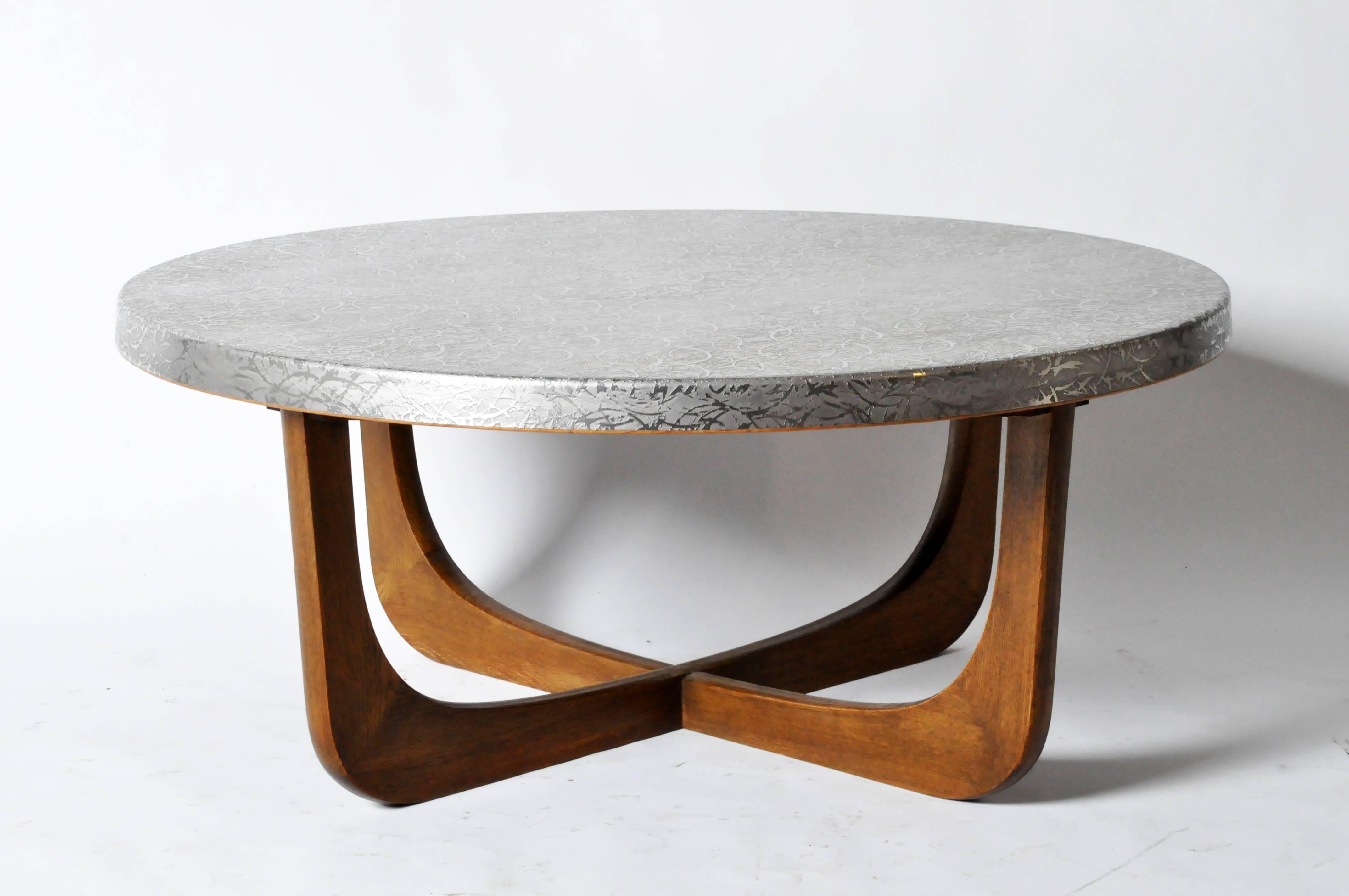 This vintage coffee table features a circular, textured aluminum top that is supported by U-shaped or ‘boomerang’ legs, which join to create an X-form base.