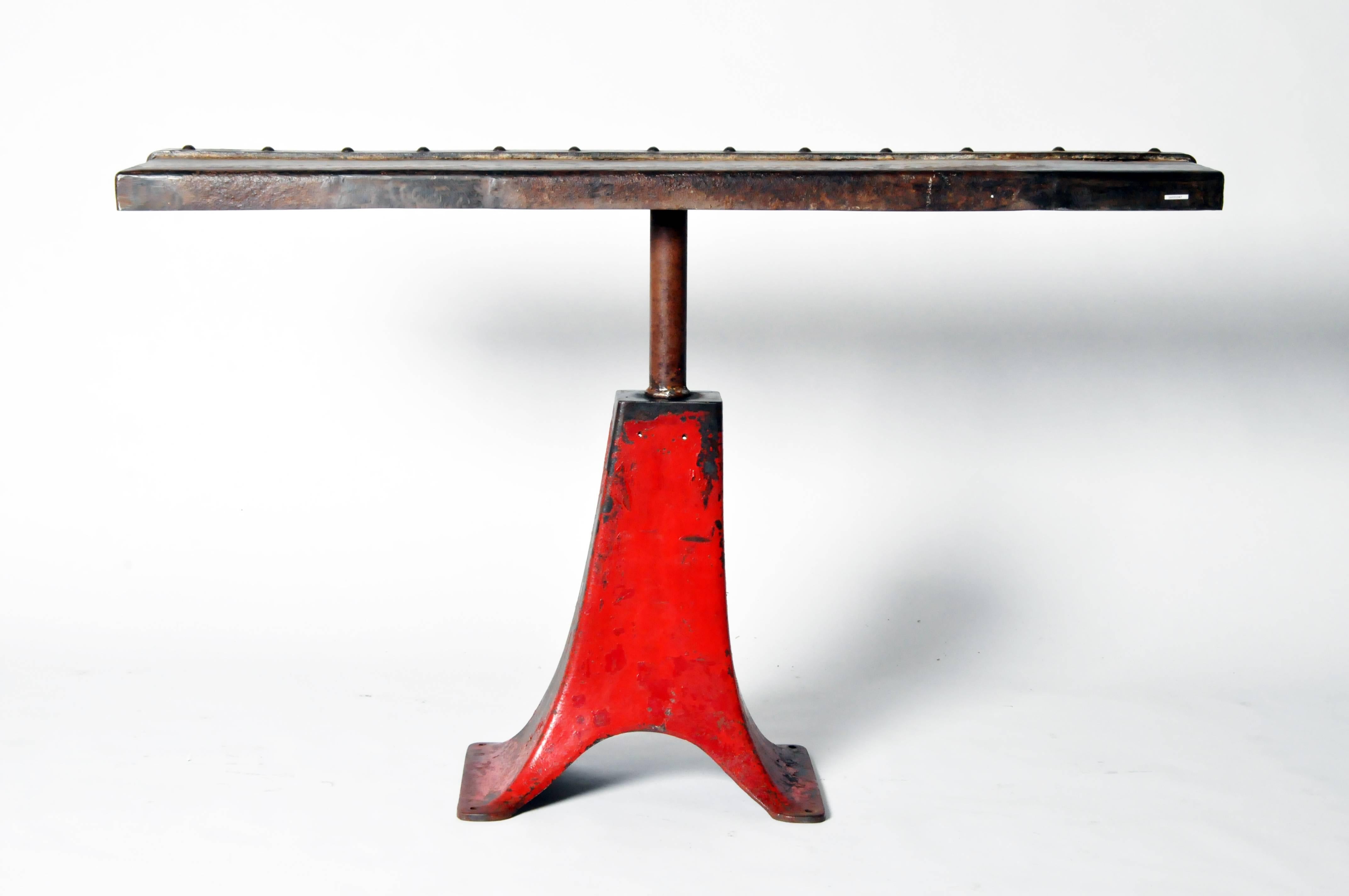 Raised on a red pedestal base, this iron table features a slab-like top with a machine riveted band along one edge.