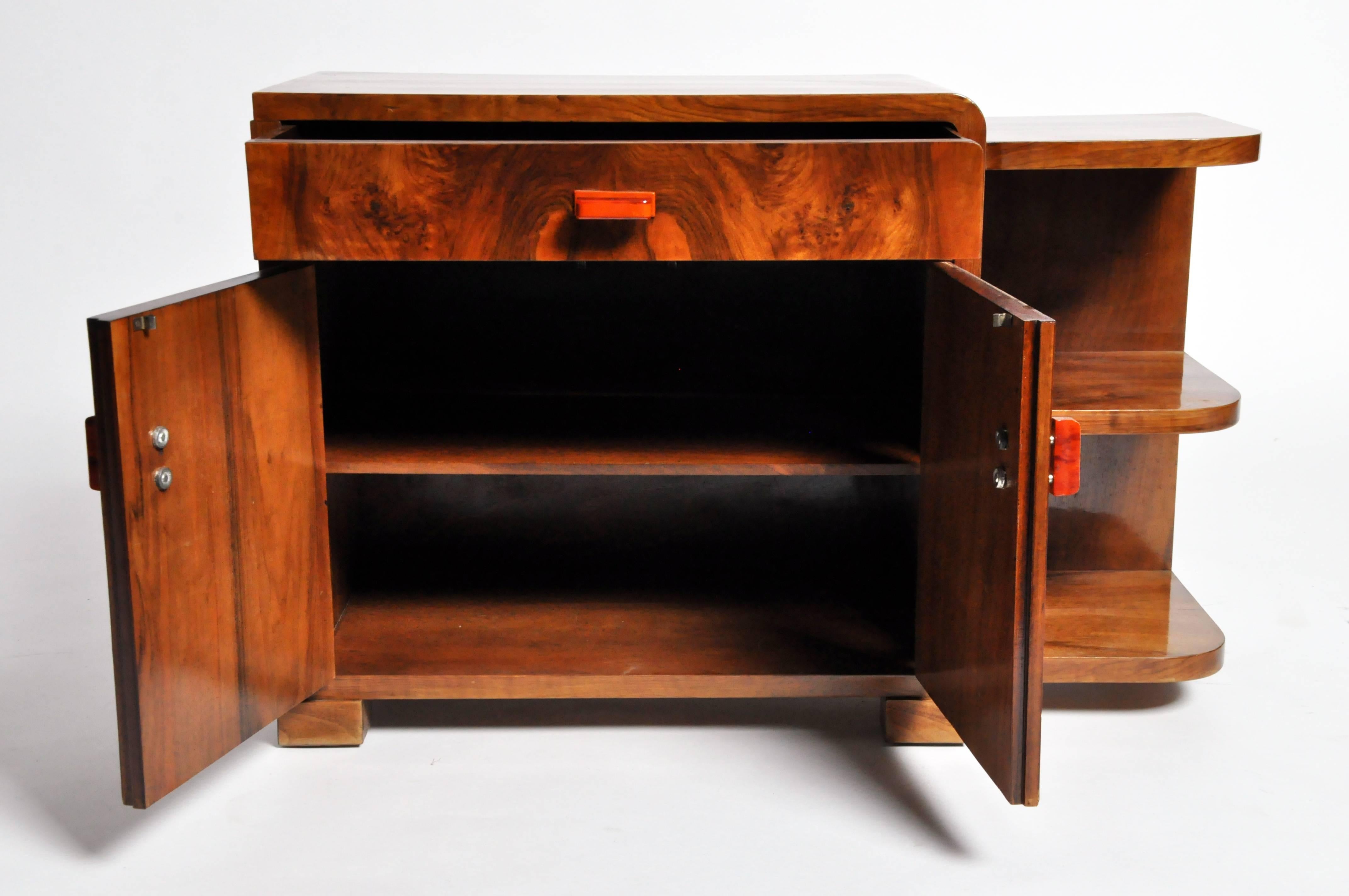 This high gloss deco side chest with drawer and shelves features stylish orange pulls and a beautiful burled walnut veneer. The sparing lines and gentle, curvilinearity of the shelves on this piece impart the sleek elegance of Europe’s historical