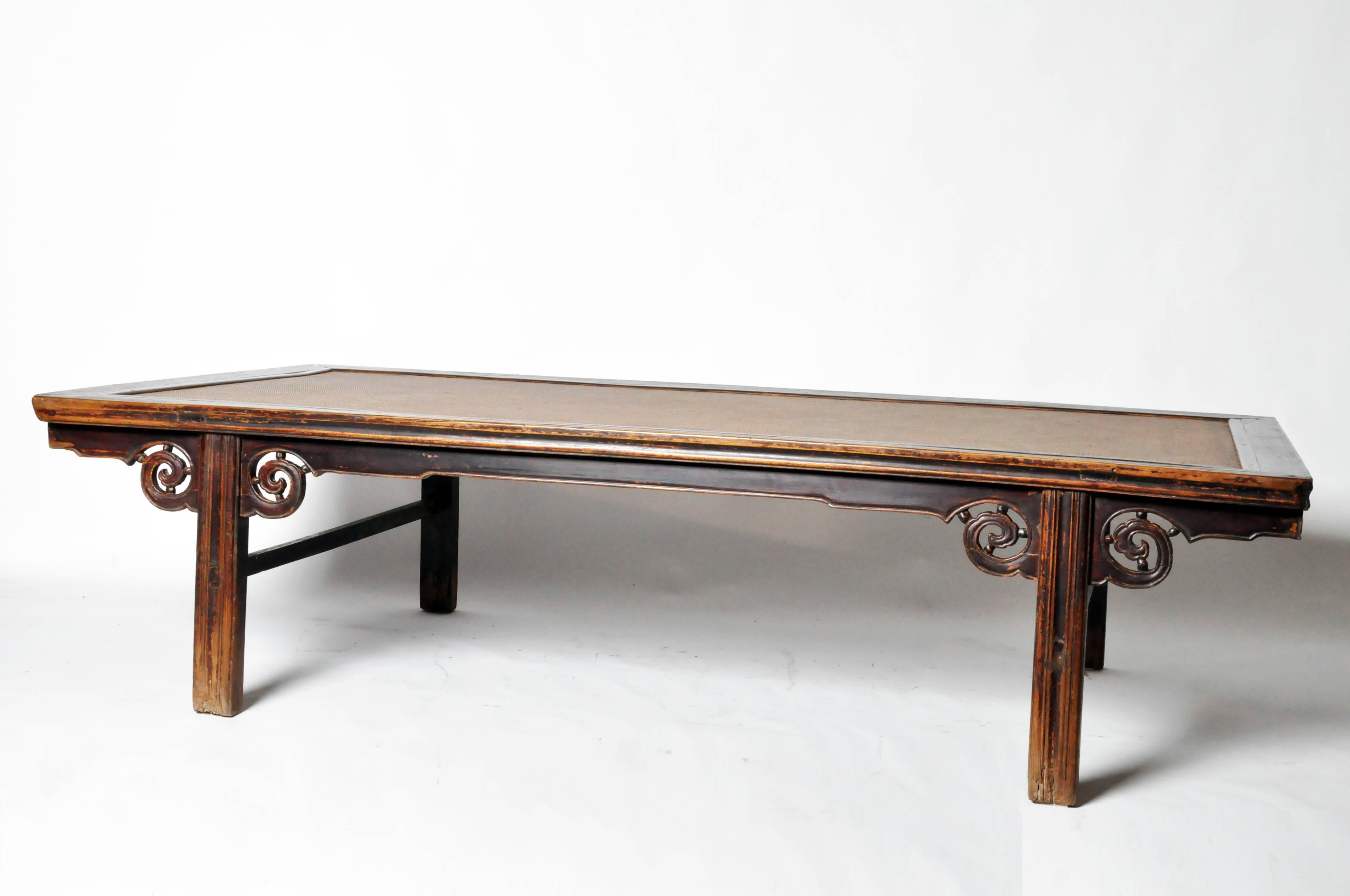 A Kang table—also referred to as a bench—is a type of Chinese furniture that serves a Dual purpose as both a low table and a chair-level bed. This particular example has a rattan top, cloud motif apron-head spandrels with beading and square legs