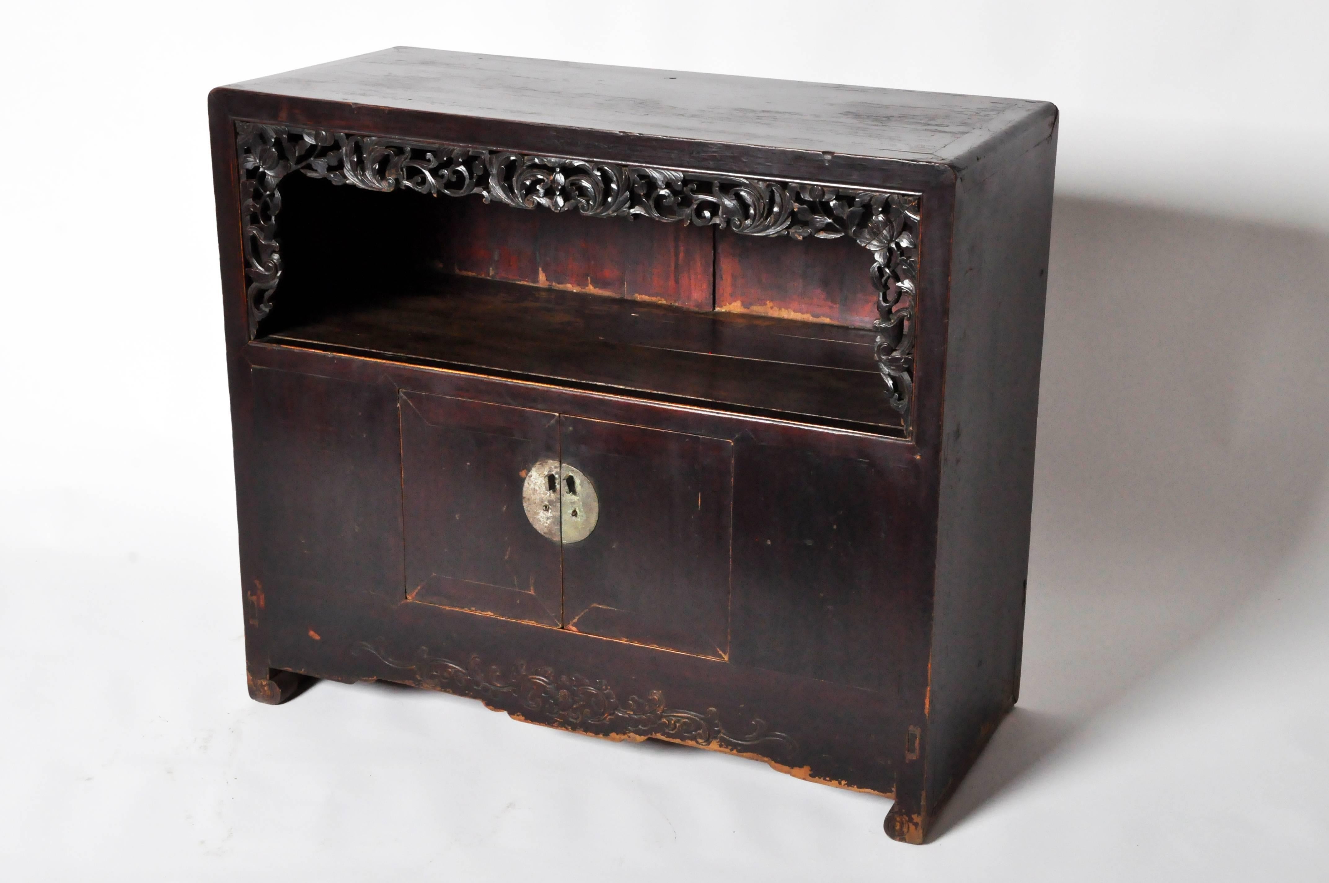 This hybrid features rounded edges on the top board, which frame a display case with an openwork foliage motif carved apron. Below the display shelf are two small doors with a circular lockplate. The foliage motif carved in relief continues at the
