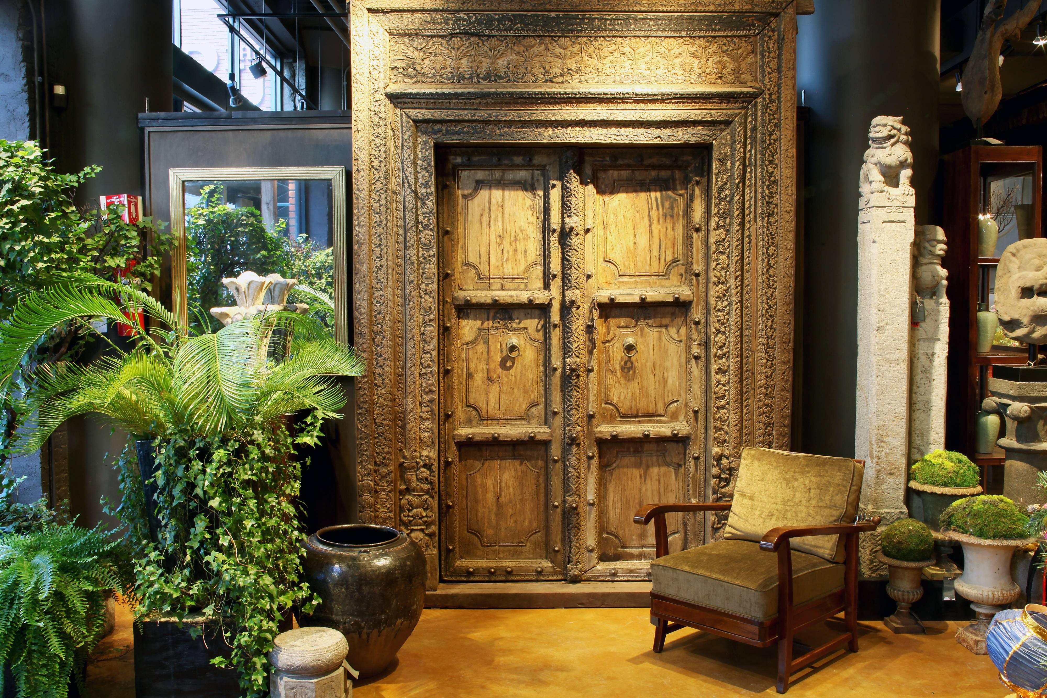 This complete entryway is handsomely relief-carved; the frame features intricate floral motifs and geometric patterns throughout. The paneled doors, with metal studs and hardware, open inwardly and can be secured with a chain and pin lock. The whole
