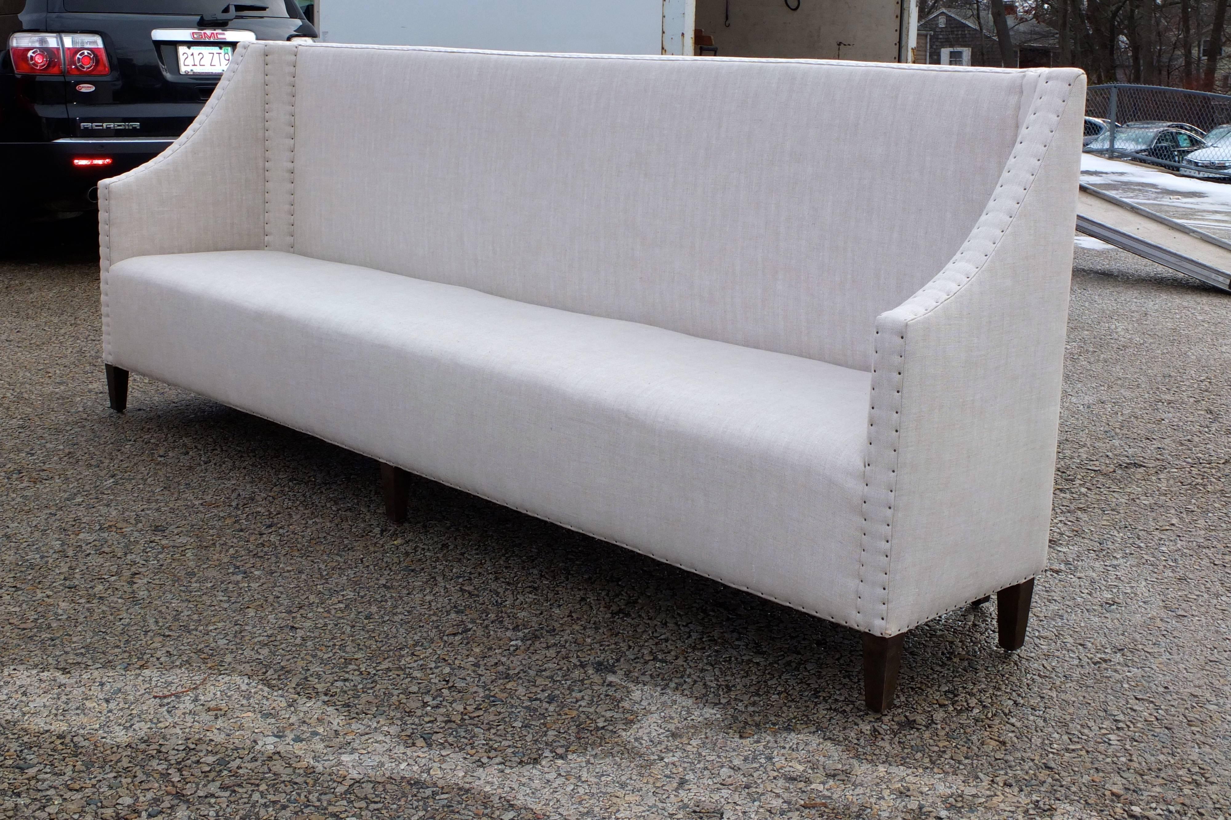 Bespoke high back banquette sofa upholstered in stone Belgian linen accented with steel nail heads and rising track arms and six tapered stained wood legs. Ten feet long, 46 inches high, 29 inches deep, 20 inch seat height. In like new condition.