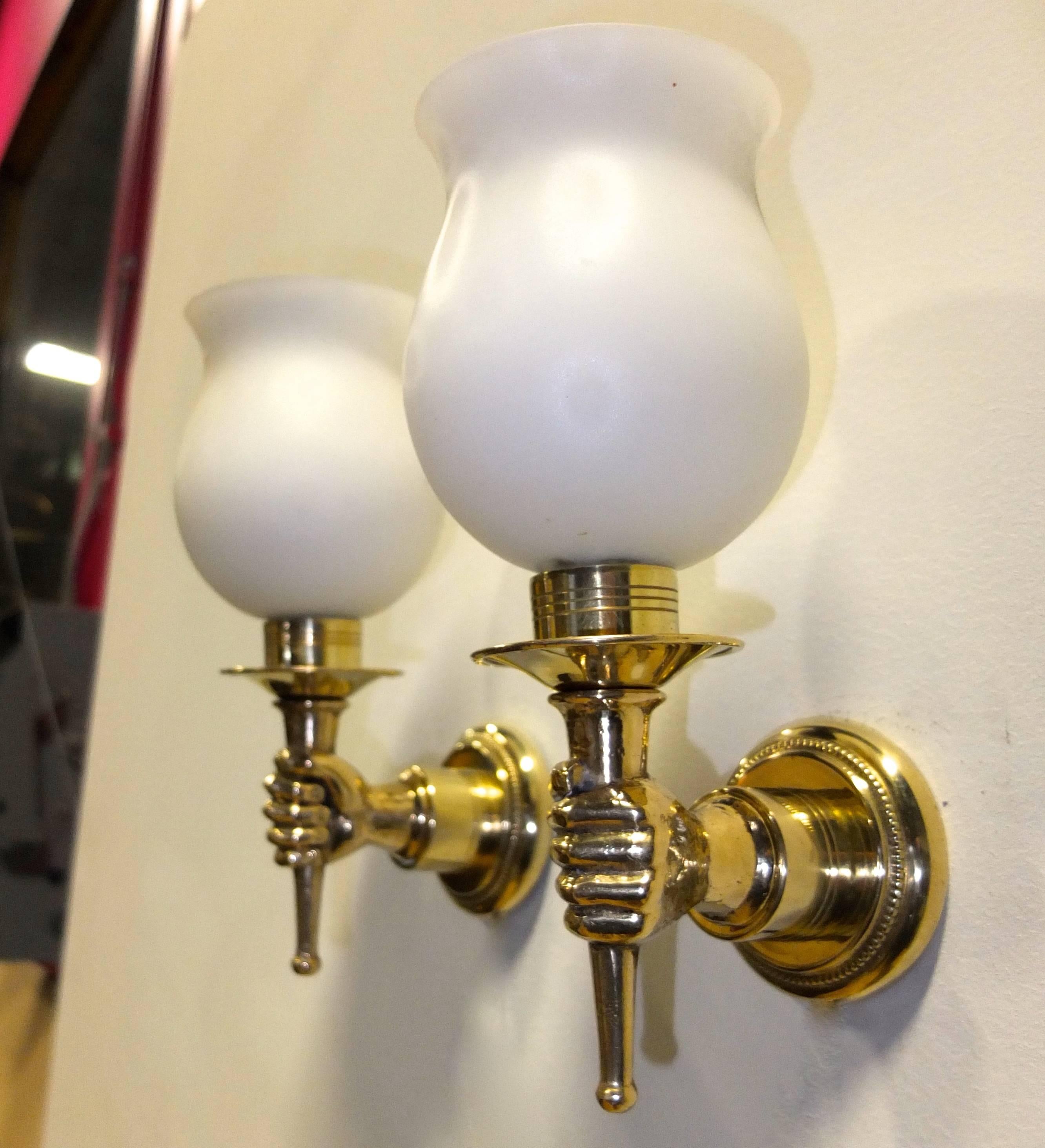 Excellent pair of French, 1950s appliques in the manner of Andre Arbus with detail of hands holding a torch made of solid and cast brass polished to a mirror finish with goblet-form opaline glass shades, rewired for USA to UL standard. Each sconce