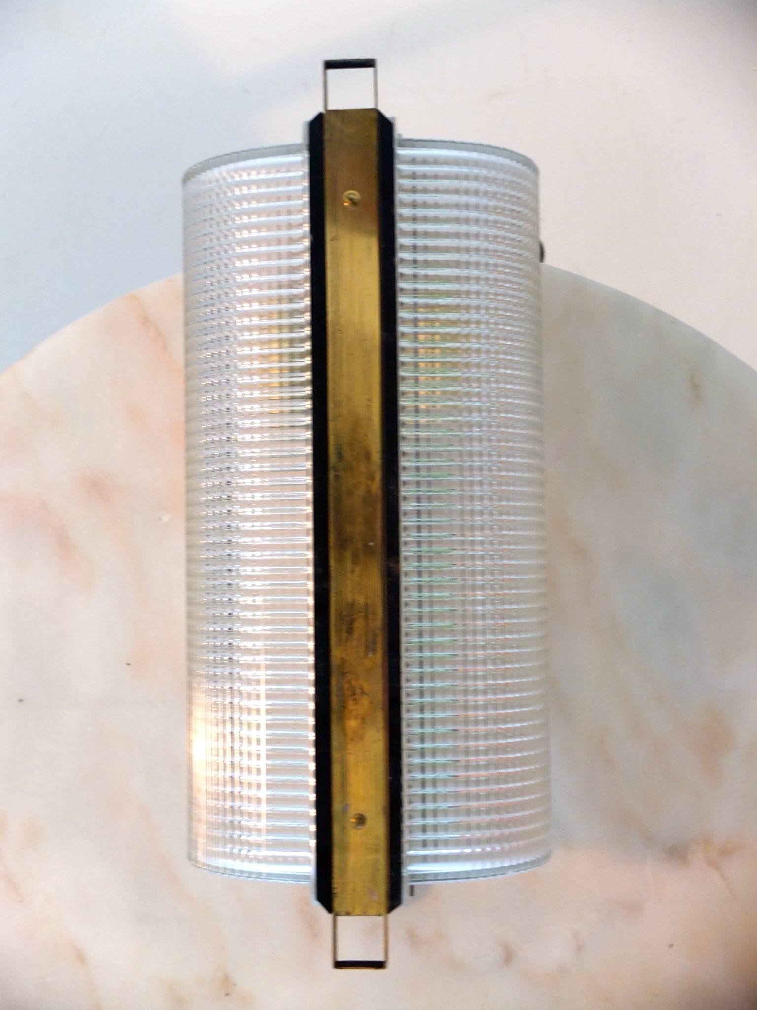 This wall mounted lamp is part of an unusual collection of lighting by Stilnovo which I acquired as a group. The glass is textured similar to Fresnel or Holophane or the lens of an automobile headlamp. The glass is in two halves banded by the matte