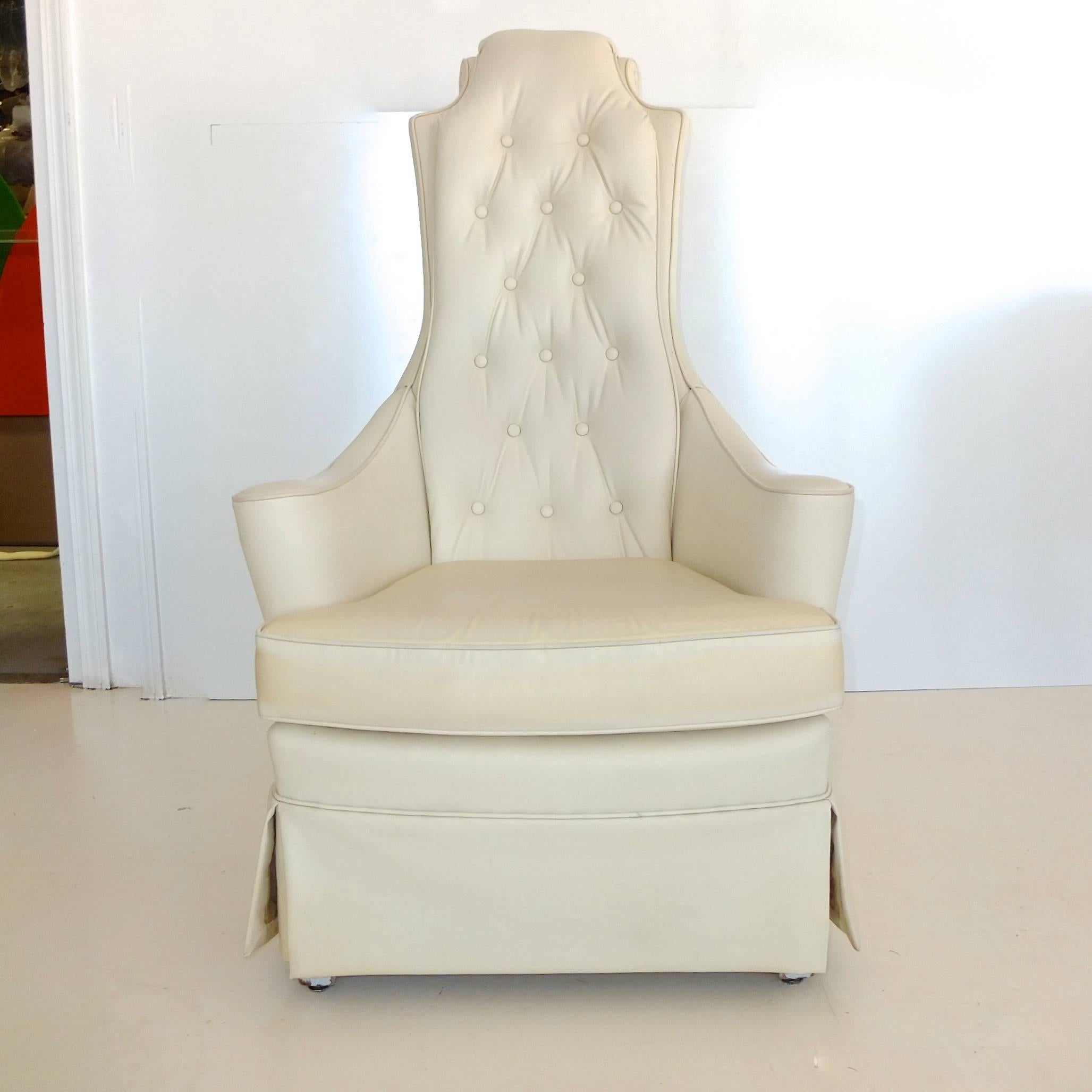 Hip kitsch Hollywood Regency high back armchair upholstered in vanilla bean naughahyde with button tufted back, loose seat cushion and pleated skirt underneath which is four painted wood legs. Lovely scrolling detail on back of headrest and