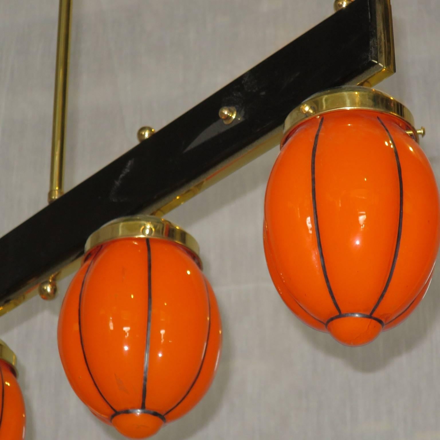 Italian midcentury colored glass chandelier,
double face black glass bar hanging by a brass tube from the ceiling.
Underneath are aligned four balls in blown transparent orange glass.
The chandelier is from the midcentury design period.
The