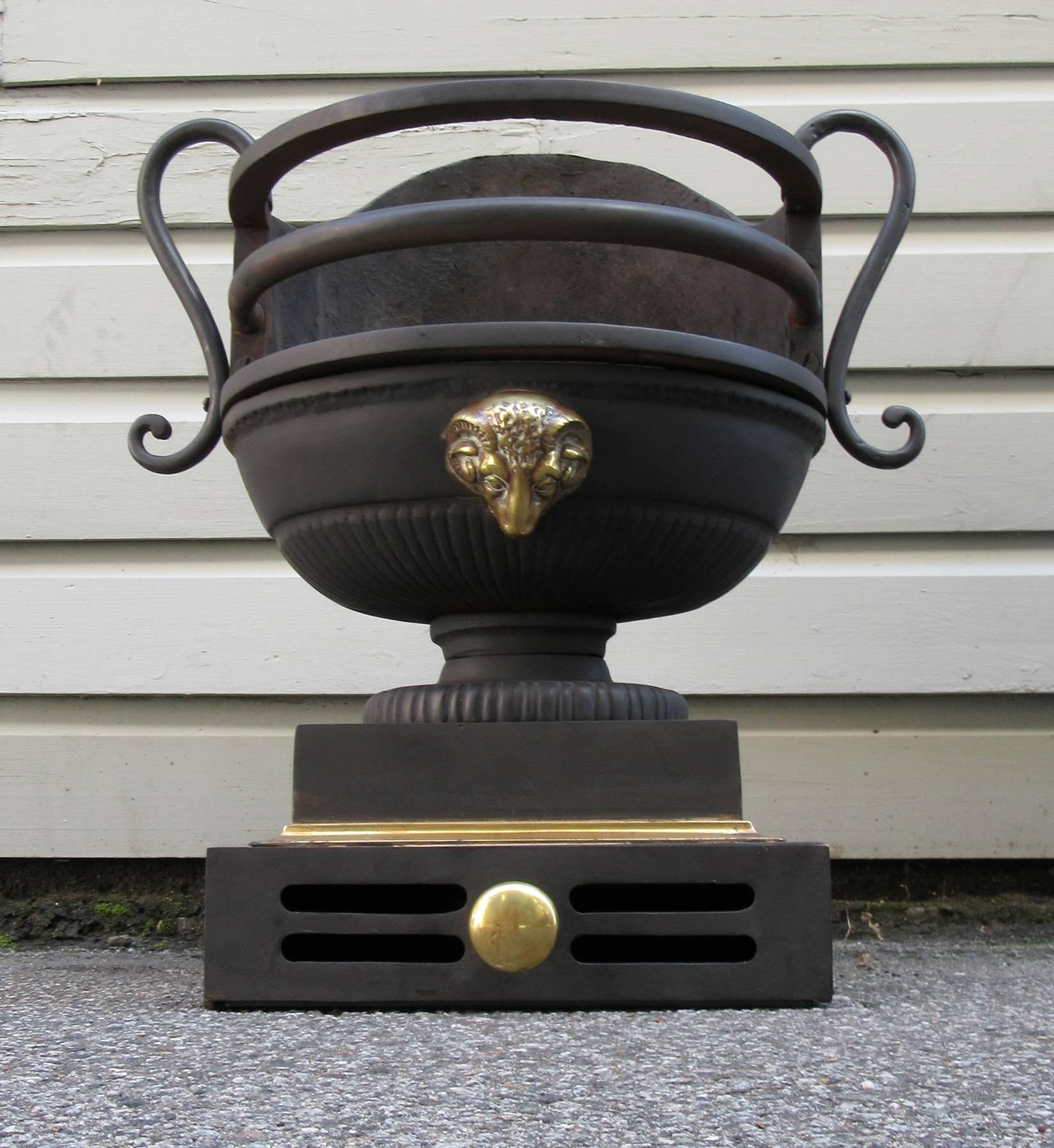An early 19th century English Regency urn shaped steel and brass fire grate, circa 1810, featuring a brass ram's head motif and ash bin with brass pull.