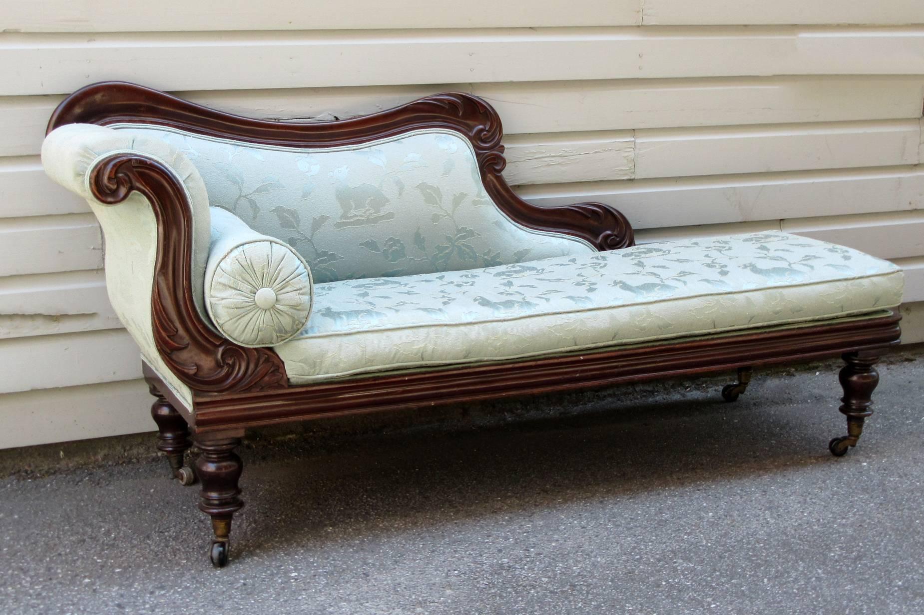 A Caribbean 19th century Jamaican Regency mahogany and upholstered recamier, circa 1830, featuring scroll arm and stylized carvings of lotus and foliage motifs. The casters are original to the chaise longue and the pale blue damask upholstery has