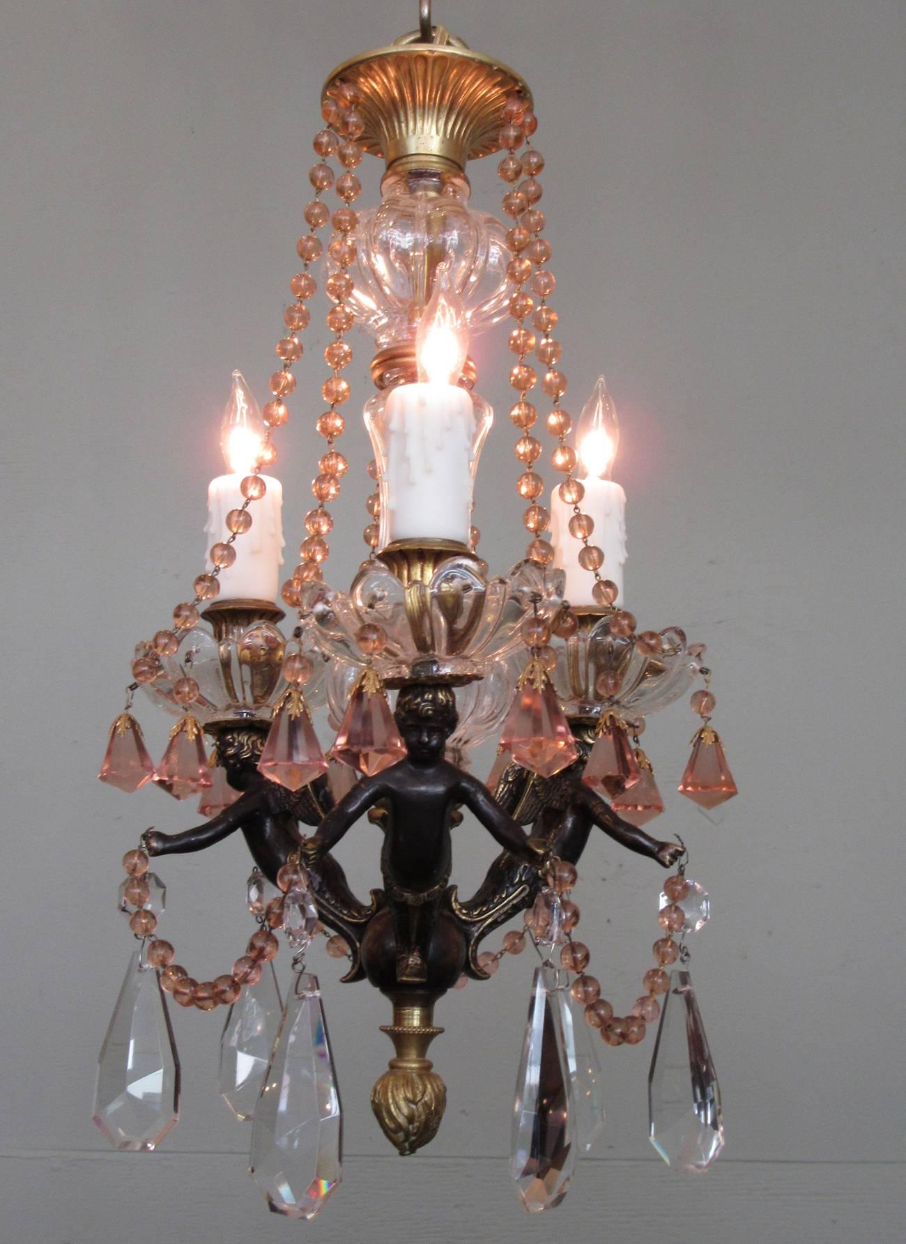 A small-scale early 20th century French Empire patinated bronze chandelier, circa 1910, featuring three candle arms adorned with putti, peach swag and crystals and clear cut crystal pendants. The chandelier has recently been cleaned and rewired with