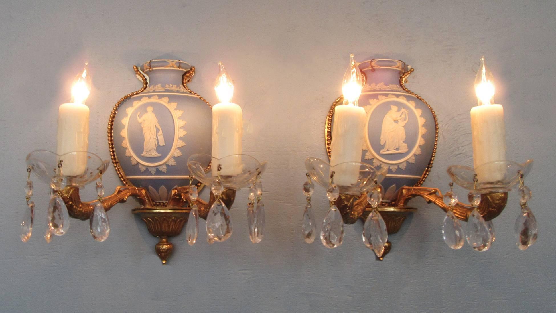 These Wedgwood sconces were made in England during the first half of the 20th century, circa 1910. These sconces feature blue urn-shaped bodies with gilded beaded edges and cameo reliefs. There are two cast-brass arms with crystal bobeches and