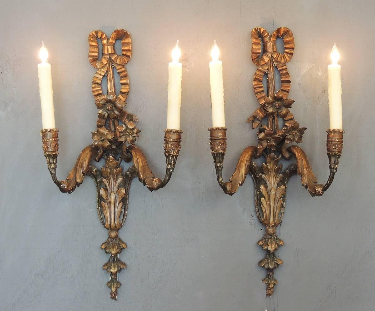 This pair of carved wood with gilt sconces were made in Italy in the early-20th century, circa 1910, and feature two candles with ribbon, flowers, and acanthus motifs. The pair has been rewired and has new porcelain sockets.