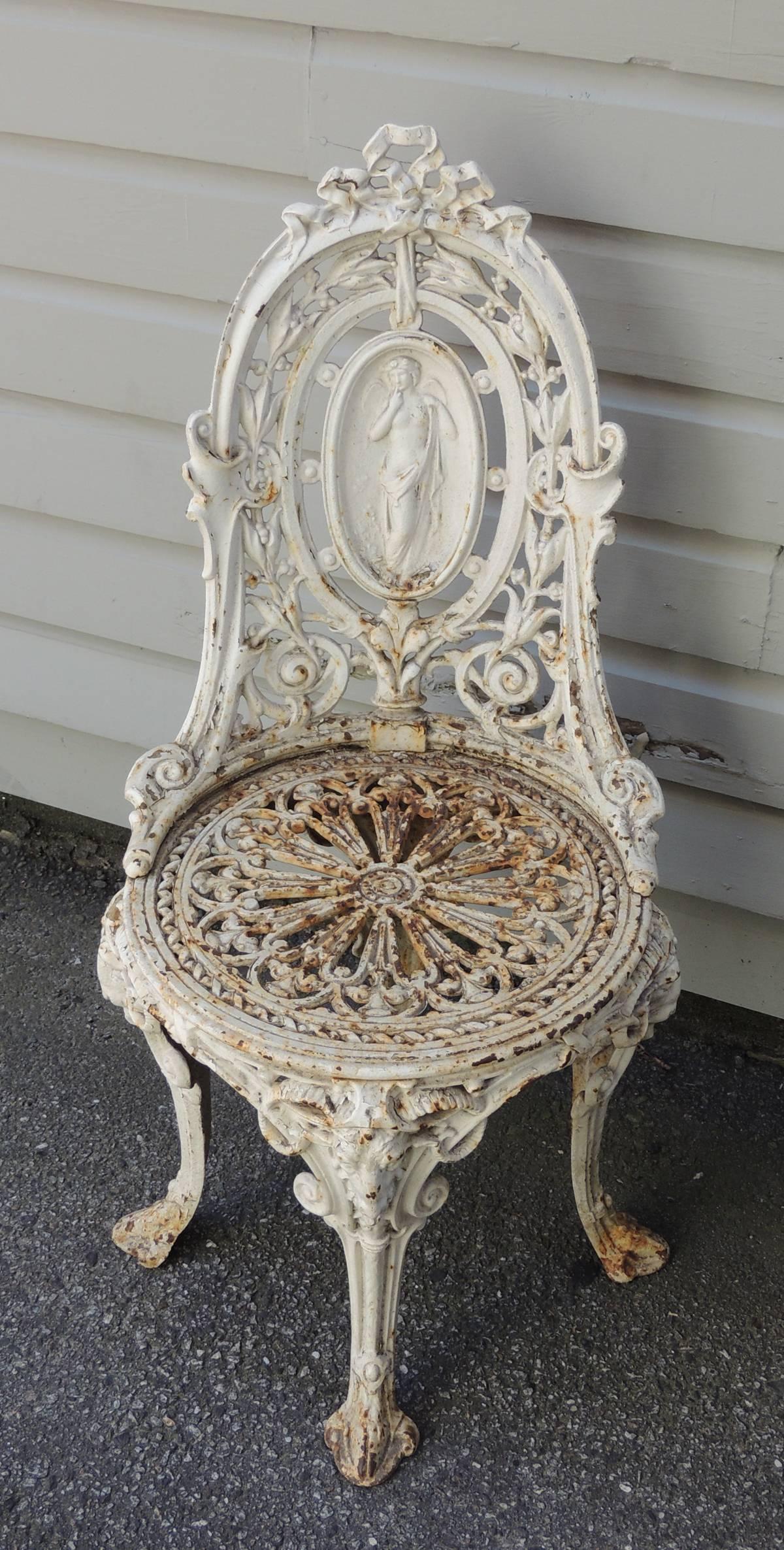This English pair of garden chairs are made of cast iron and feature classical Greek figures surrounded with scroll designs and ribbon motifs. The seats are decorated with pierced design and the chairs four legs each feature a ram's heads motif.