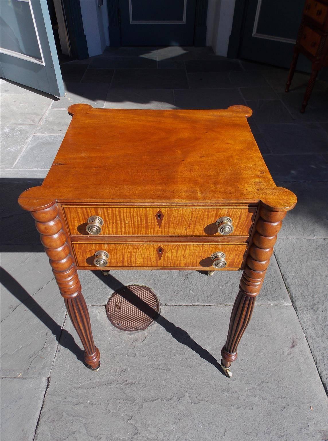 American mahogany & tiger maple two drawer stand with carved outset corners, original brass knobs, diamond inlaid escutcheons, and terminating on turned bulbous reeded legs with the original brass casters. Early 19th Century. Table is finished on