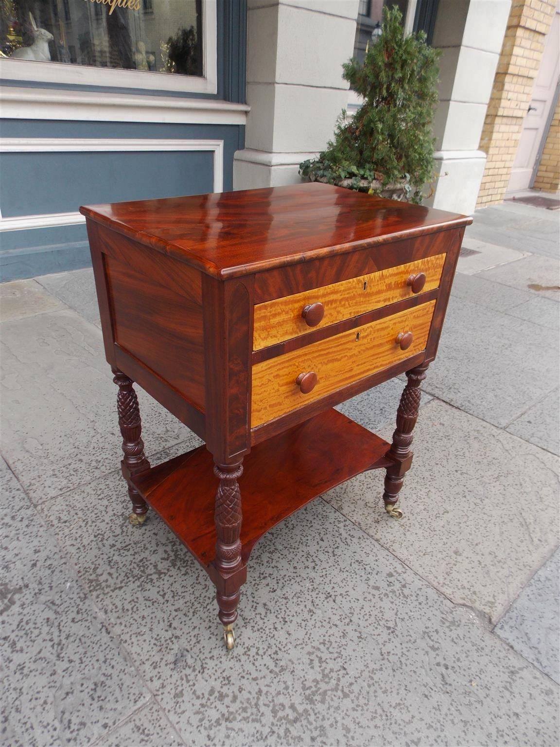 American mahogany and satinwood graduated two-drawer stand with the original wooden knobs, recessed side panels, pineapple and turned ringed legs, lower connecting shelf and terminating on original brass casters, Early 19th Century. Stand is
