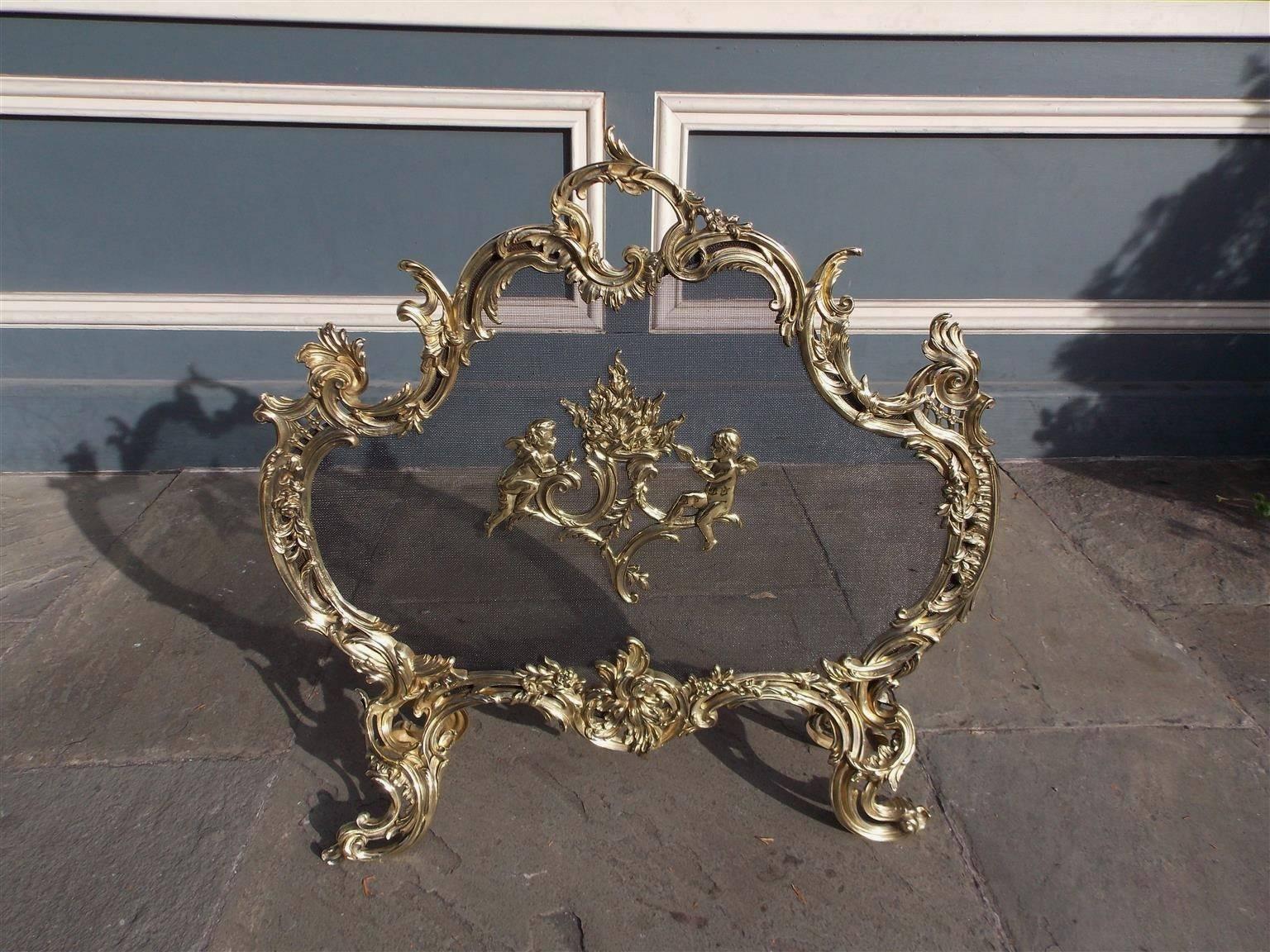 French brass free standing fire screen with flanking cherubs tending to a flame medallion, centered floral acanthus handle, scrolled filigree sides, and terminating on scrolled floral decorative motif feet.  Early 19th Century.  