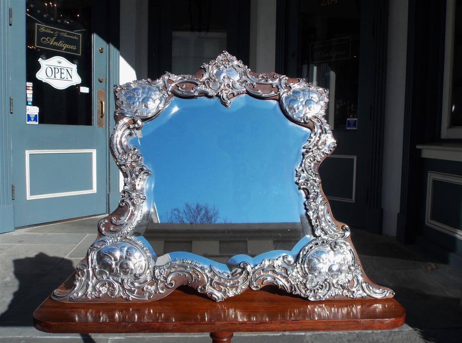 Specchio da toeletta inglese in argento massiccio con cartiglio floreale centrale, quattro cherubini alati angolari affiancati, lati decorativi floreali a volute, marchi di fabbrica e poggiato su una cornice angolare in legno intagliato. Lo specchio