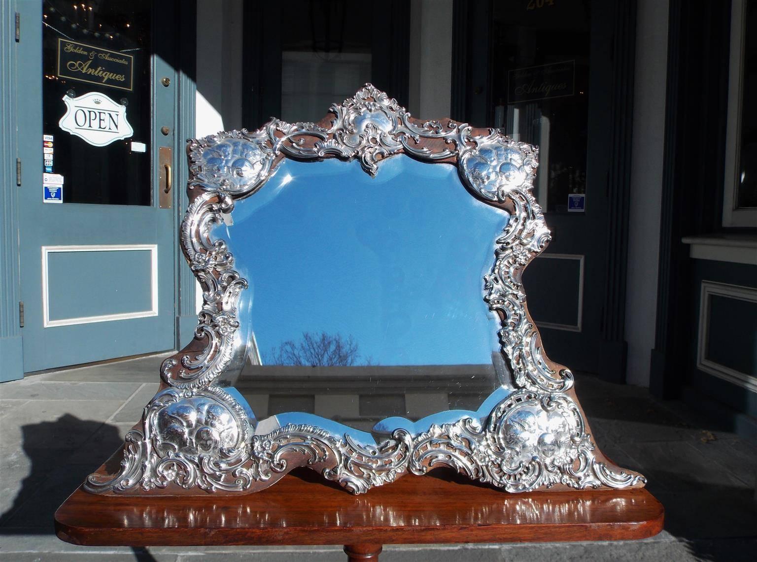 Miroir de toilette anglais en argent sterling avec cartouche floral au centre, quatre chérubins ailés dans les coins, côtés décoratifs floraux à volutes, poinçons, et reposant sur un cadre en bois sculpté dans les coins. Le miroir a le verre