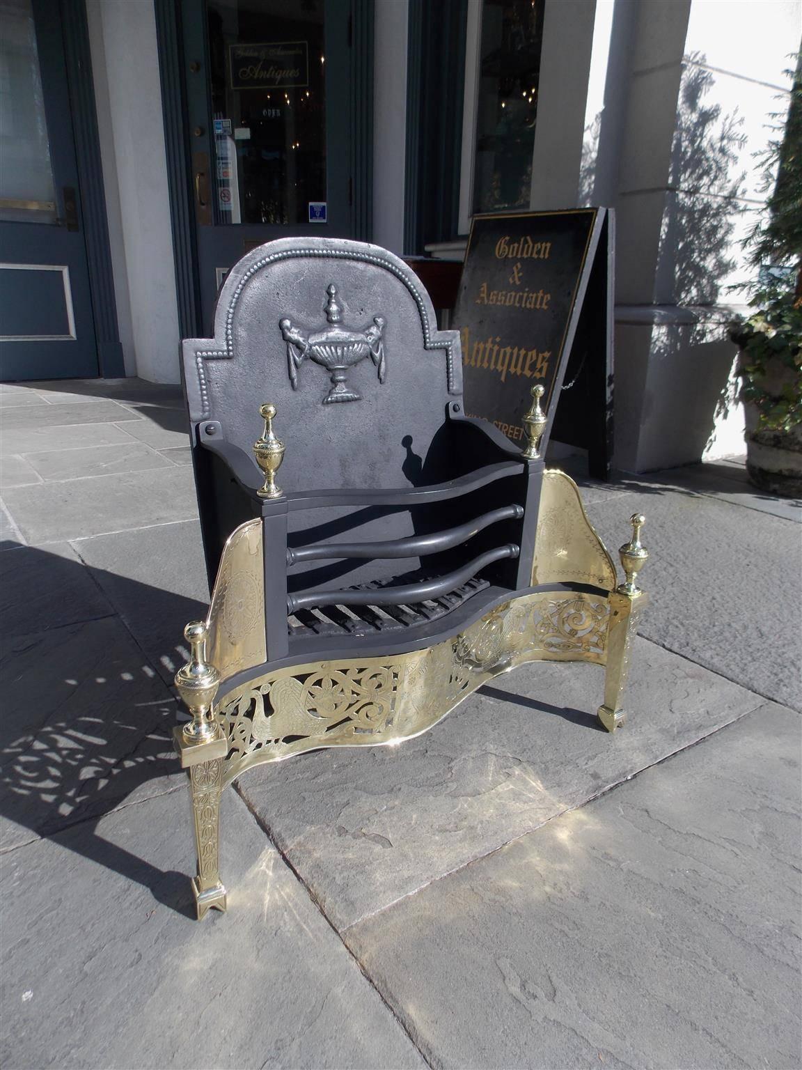 English cast iron and brass serpentine coal grate with tombstone back, centered Adam urn, flanking brass finials, coal basket, hand chased skirt with flanking cranes, centered urn, decorative floral motif and medallions with graduated bell flowers,