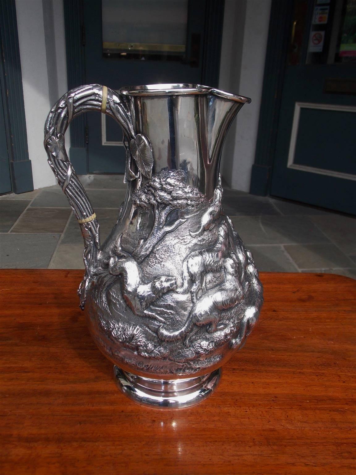 English sterling silver pitcher with embossed hunting scenes depicting a gentleman with his canine companions spearing an otter from a stream with a vine and ivory foliage motif handle all terminating on a circular ribbed base. Late 18th Century. 
