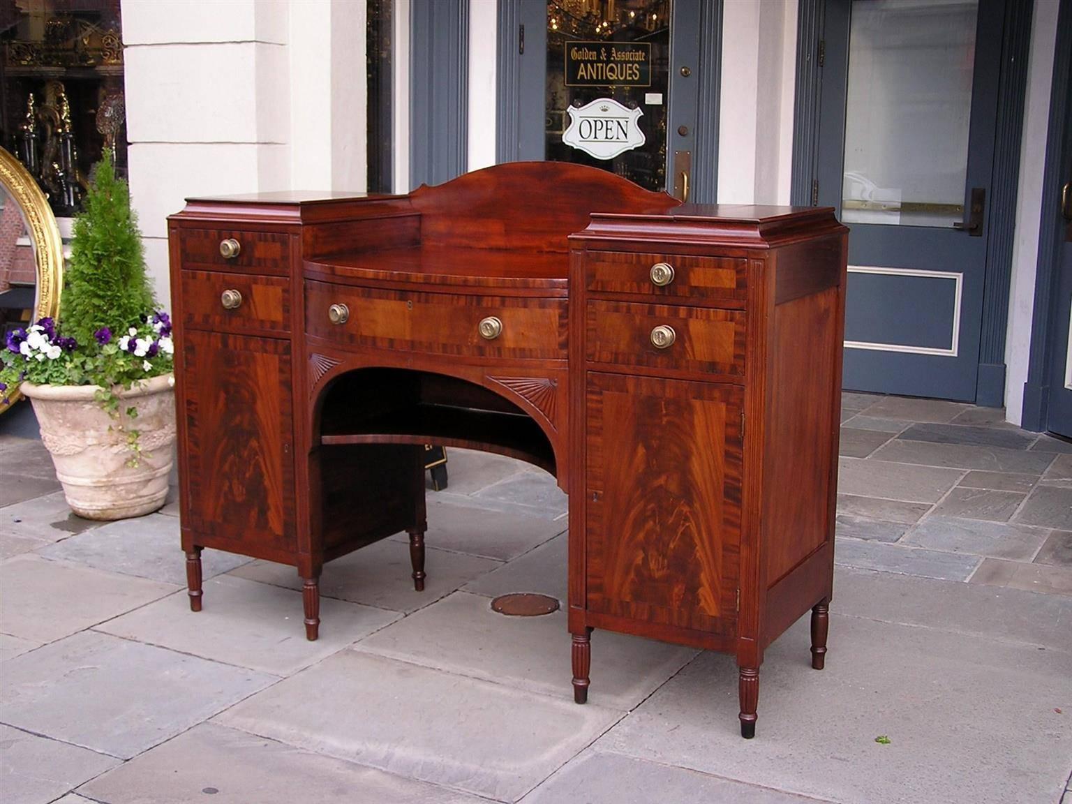 American Sheraton mahogany sideboard with a centered splash board, flanking cross banded cabinets containing five fitted drawers with the original brasses, carved corner fan motif, lower shelf, and terminating on reeded turned ringed legs. Early