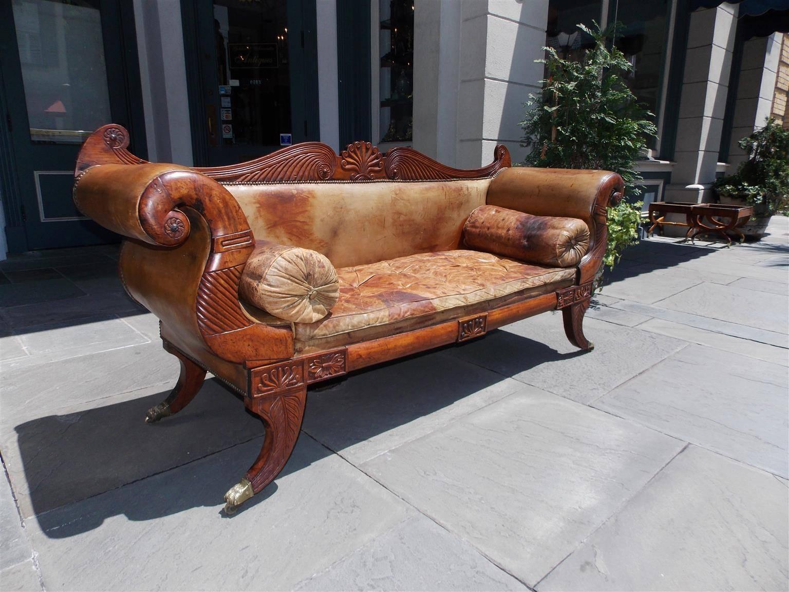 Caribbean Regency mahogany leather sofa with a central serpentine and scrolled fluted back, central floral shell with flanking scrolled medallions, flanking fluted carved arms with bolster side pillows, carved molded edge foliage skirt, and resting