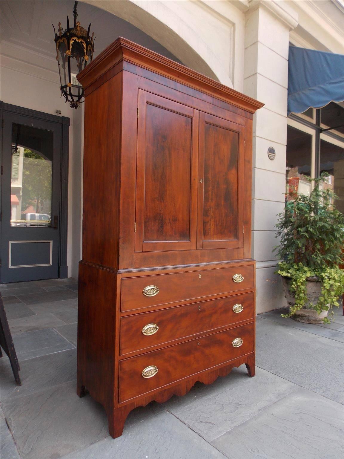American cherry & mahogany linen press with carved molded edge cornice, upper case flanking book matched doors revealing three interior shelves. Lower case has three graduated drawers with period brasses, and terminating with a carved scalloped