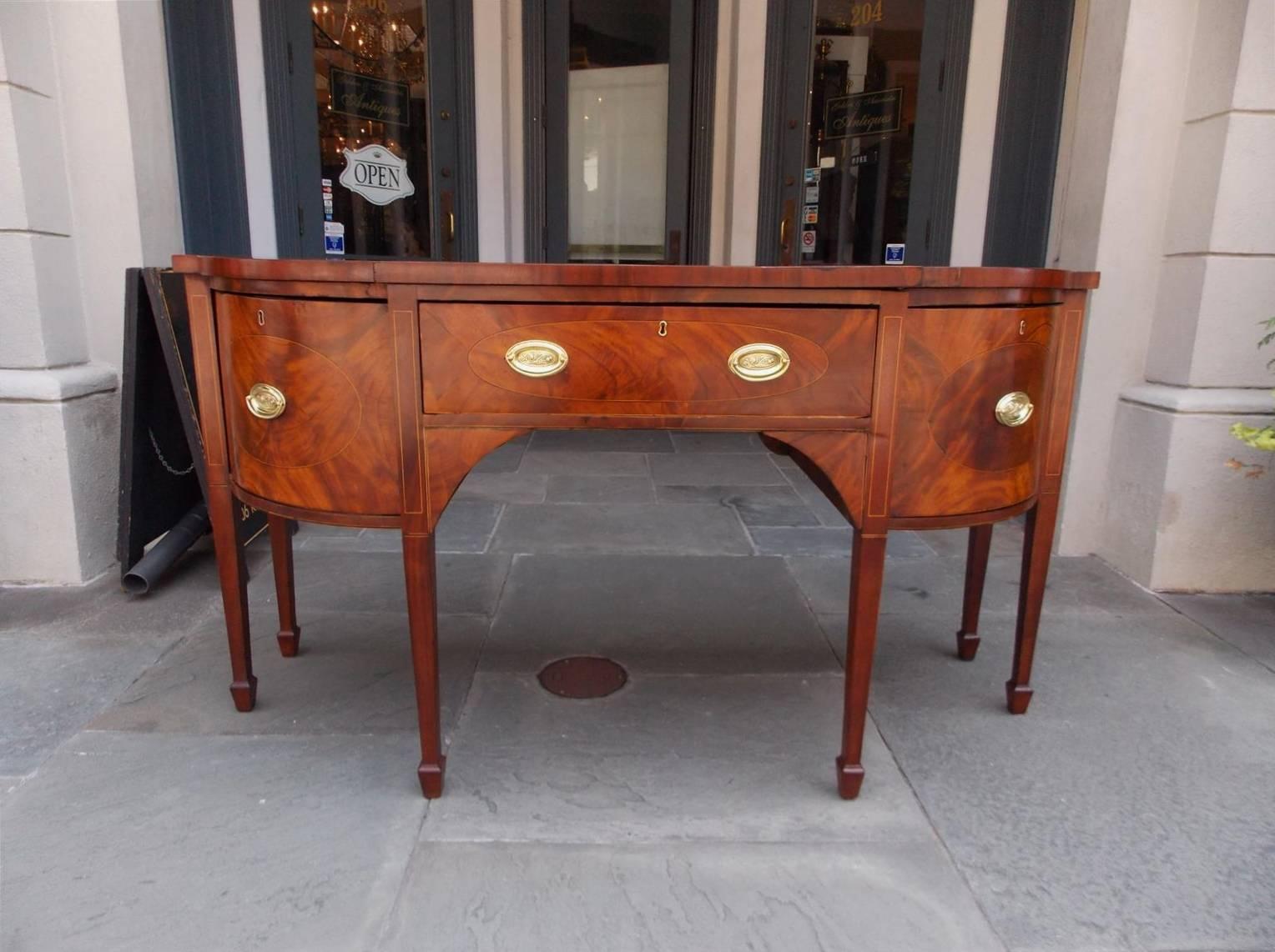 English Hepplewhite mahogany bow front cross banded sideboard with a carved molded edge top, central drawer with flanking cabinet doors, oval satinwood string inlays,  period brasses, and terminating on tapered squared legs with spade feet, Late