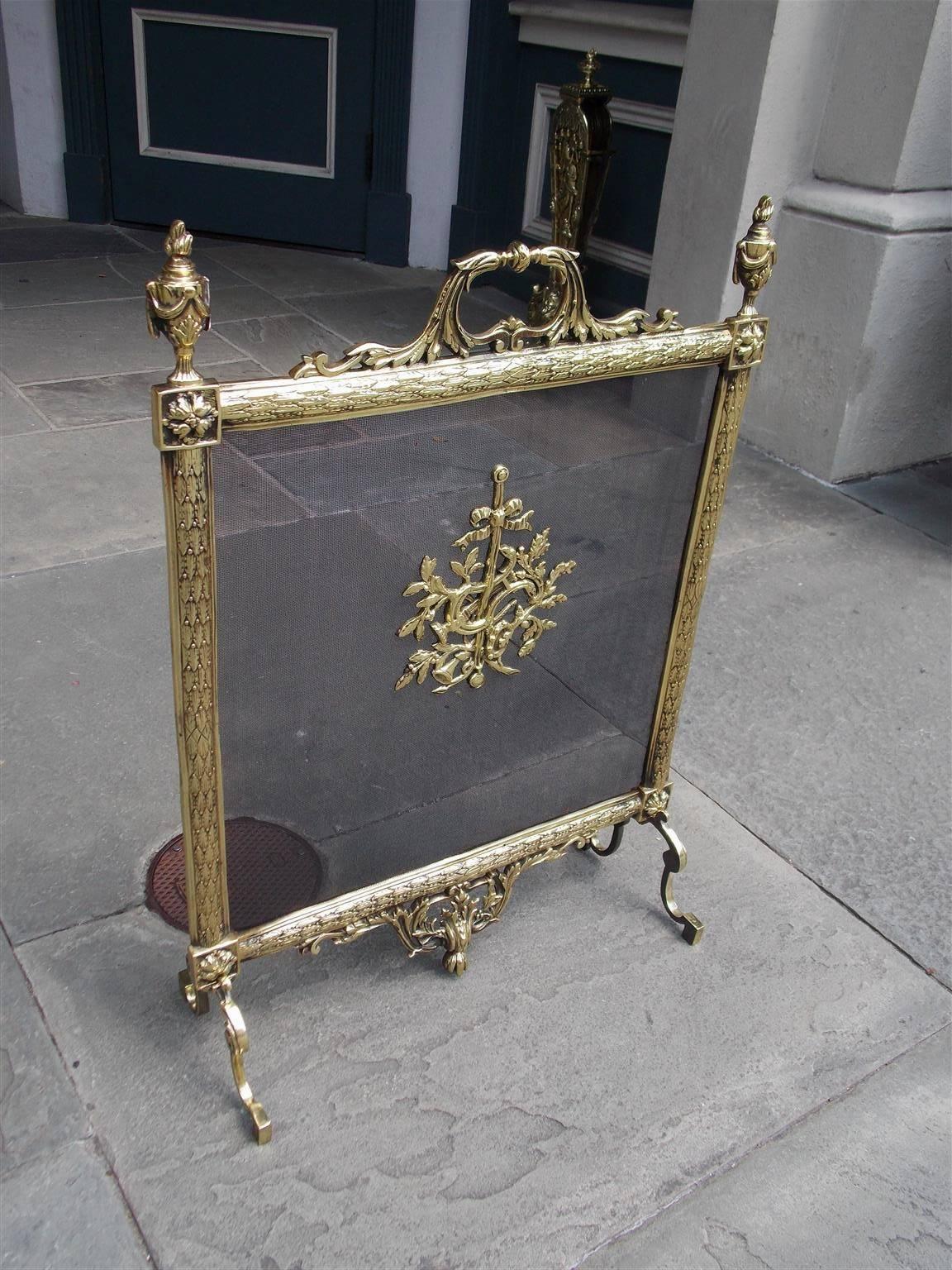 French brass freestanding fire screen with acanthus scrolled centered handle, flanking flame urn swag finials, corner floral medallions, centered decorative ribbon with floral lyre motif, and terminating on flanking scrolled legs with a lower