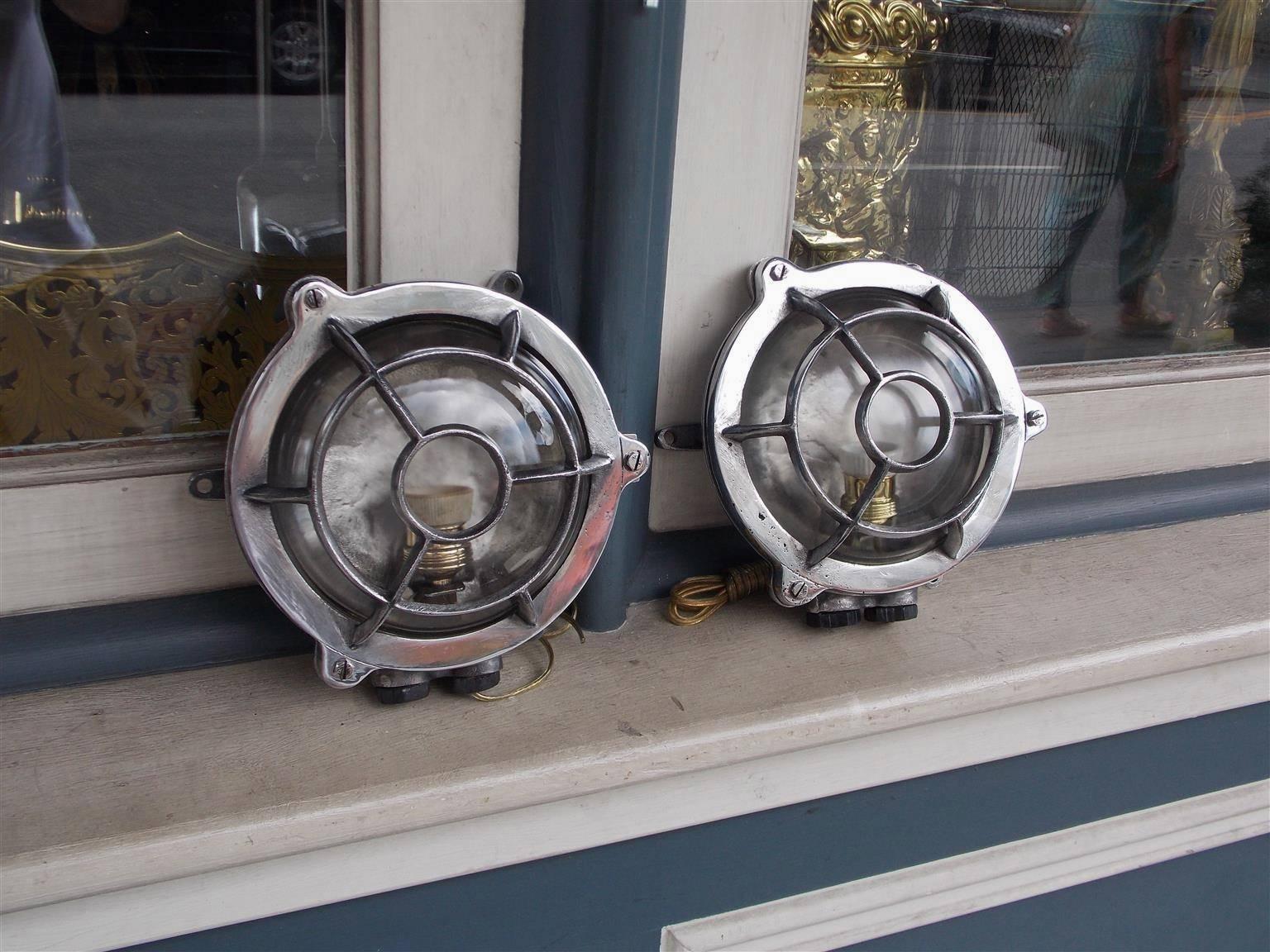 Pair of American polished aluminum bulls eye nautical wall lights with protective front cages and rear mounting brackets. Pair has been electrified with a single socket for bulb, 20th century.